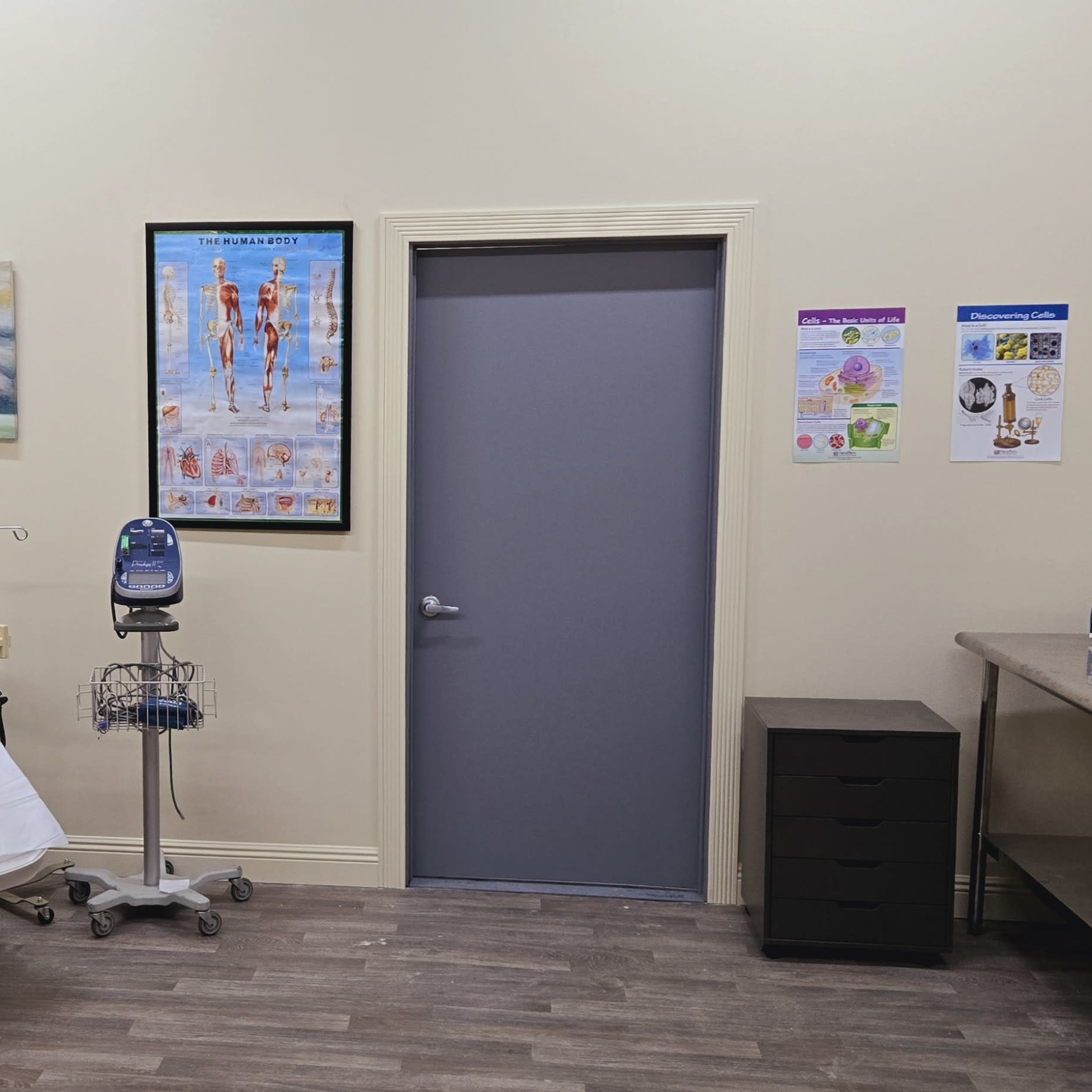A clinical examination room featuring an adjustable examination table, a rolling cart, a desk, and various posters on the walls.