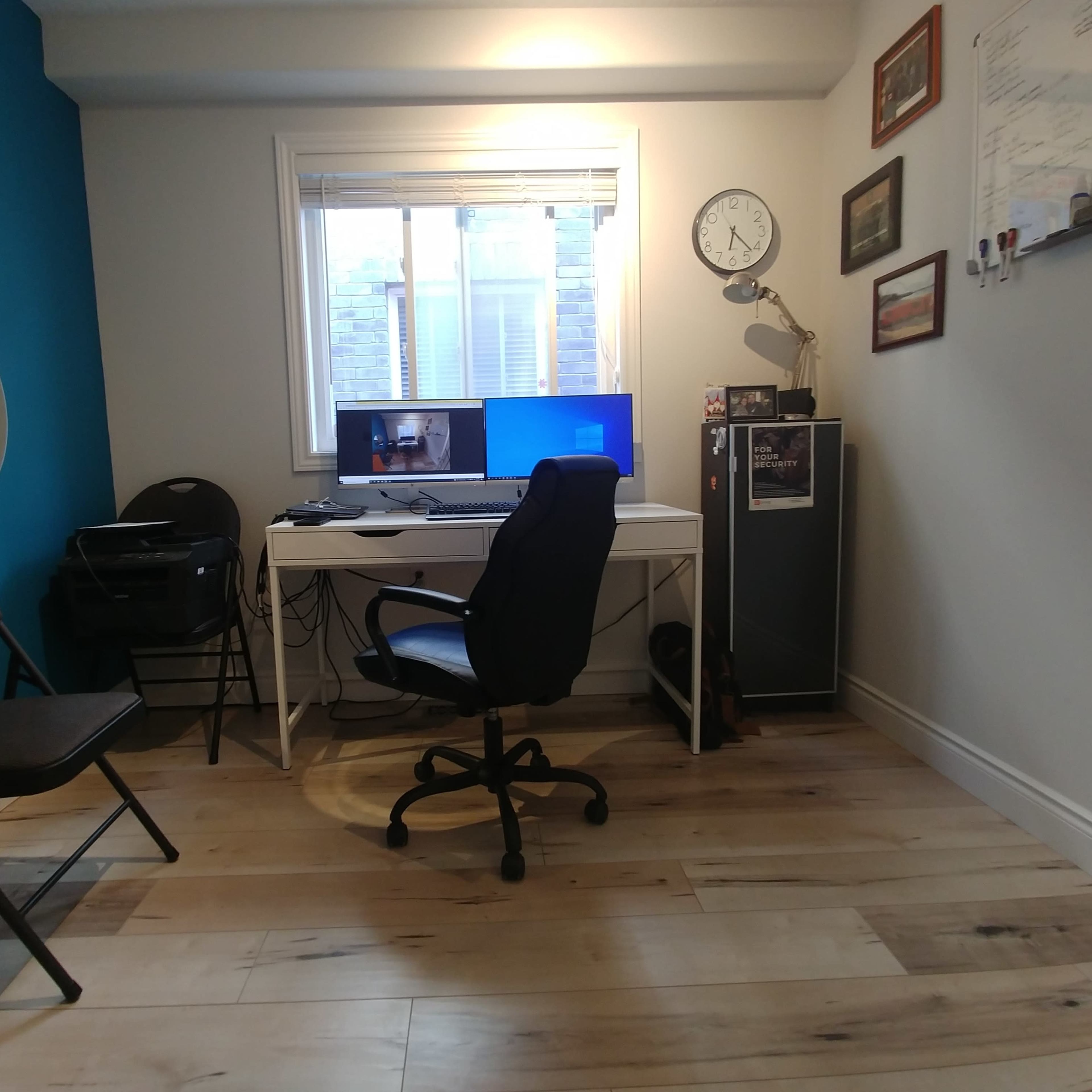 A home office is set up with a desk, computer, chair, printer, and a clock in a room with blue and white walls.