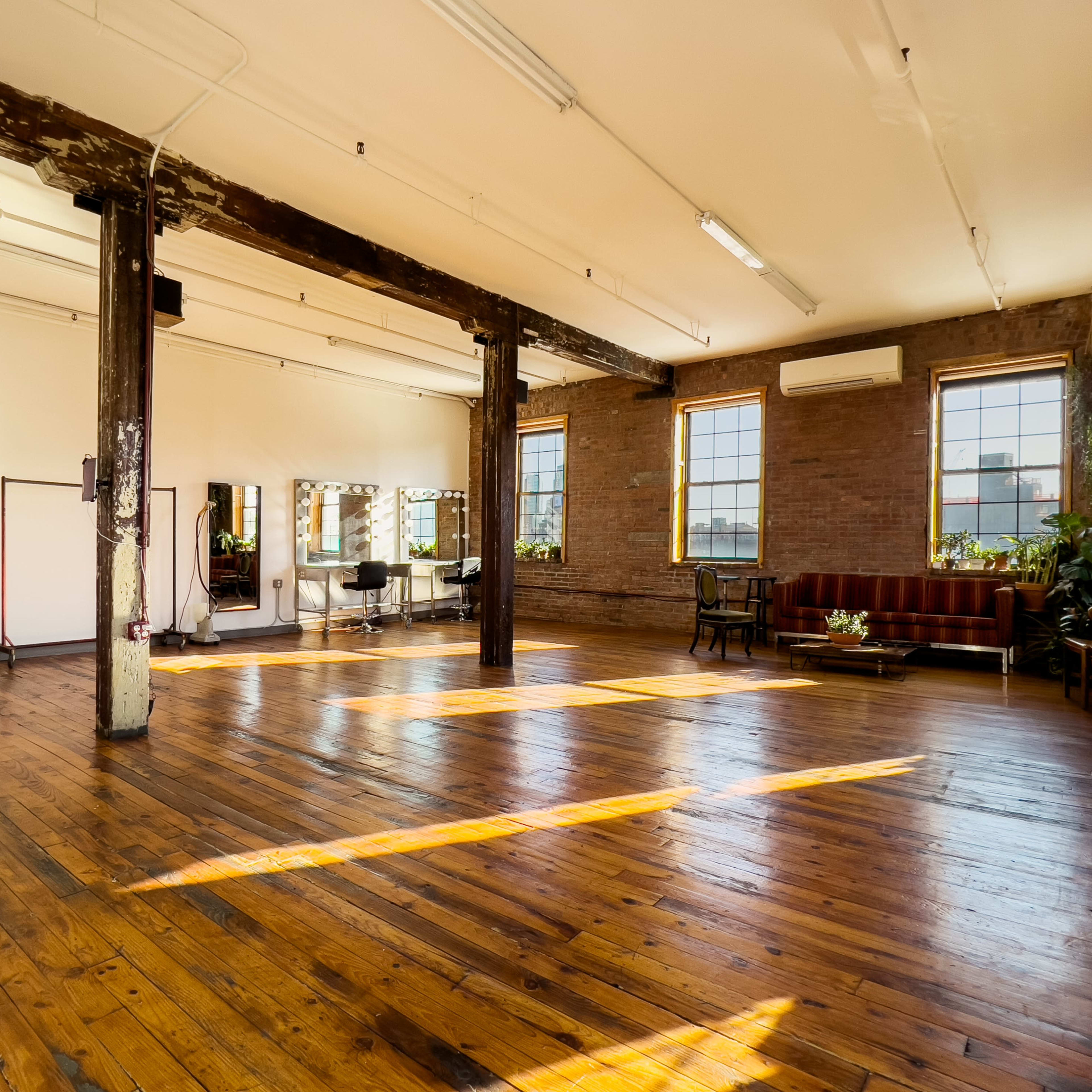 A spacious, well-lit room with wooden floors, brick walls, and large windows, featuring a few furniture pieces and mirrors.