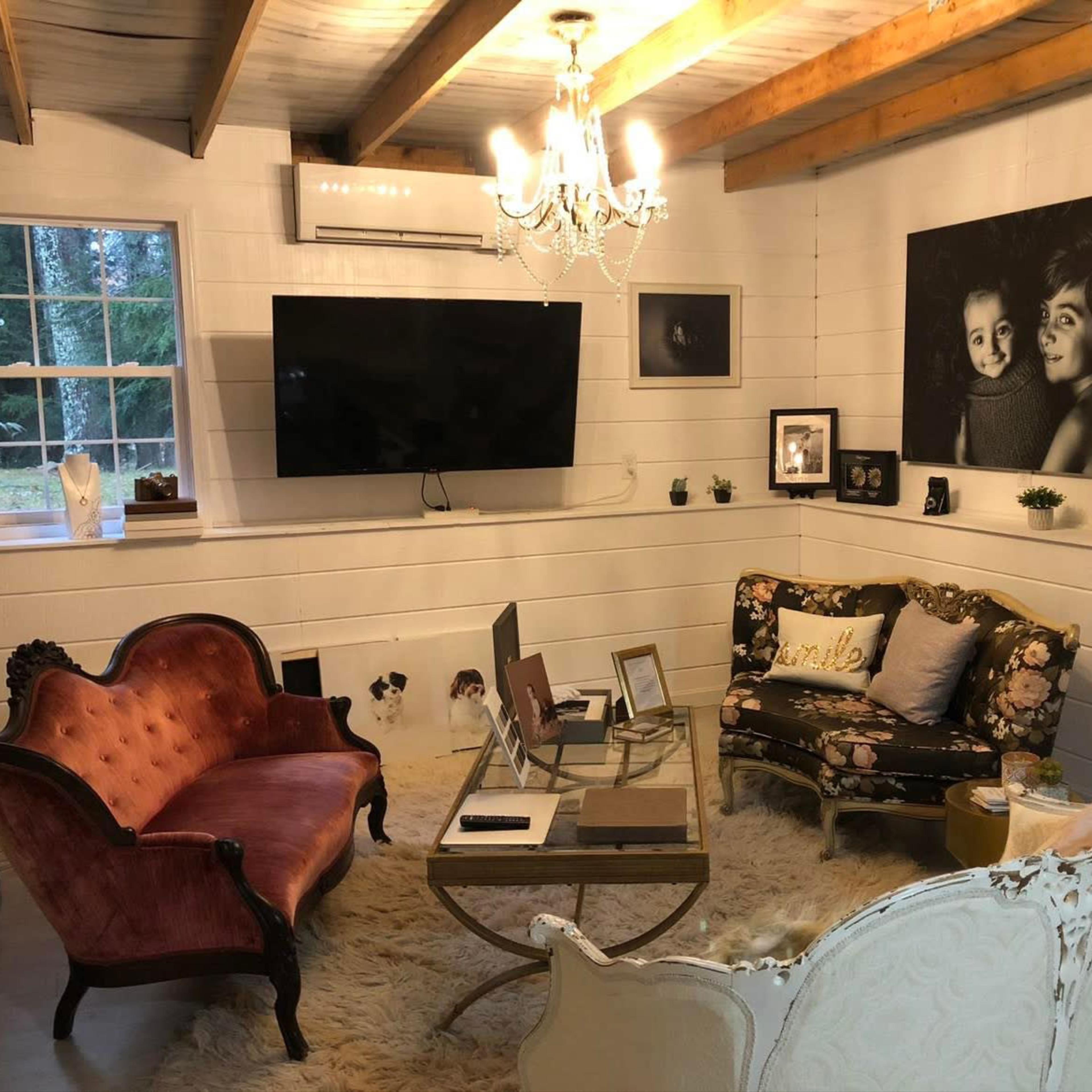 A cozy living area featuring a blend of vintage and modern furniture, including a pink velvet sofa and a floral-patterned couch, with a large TV and framed photos on the white shiplap wall.