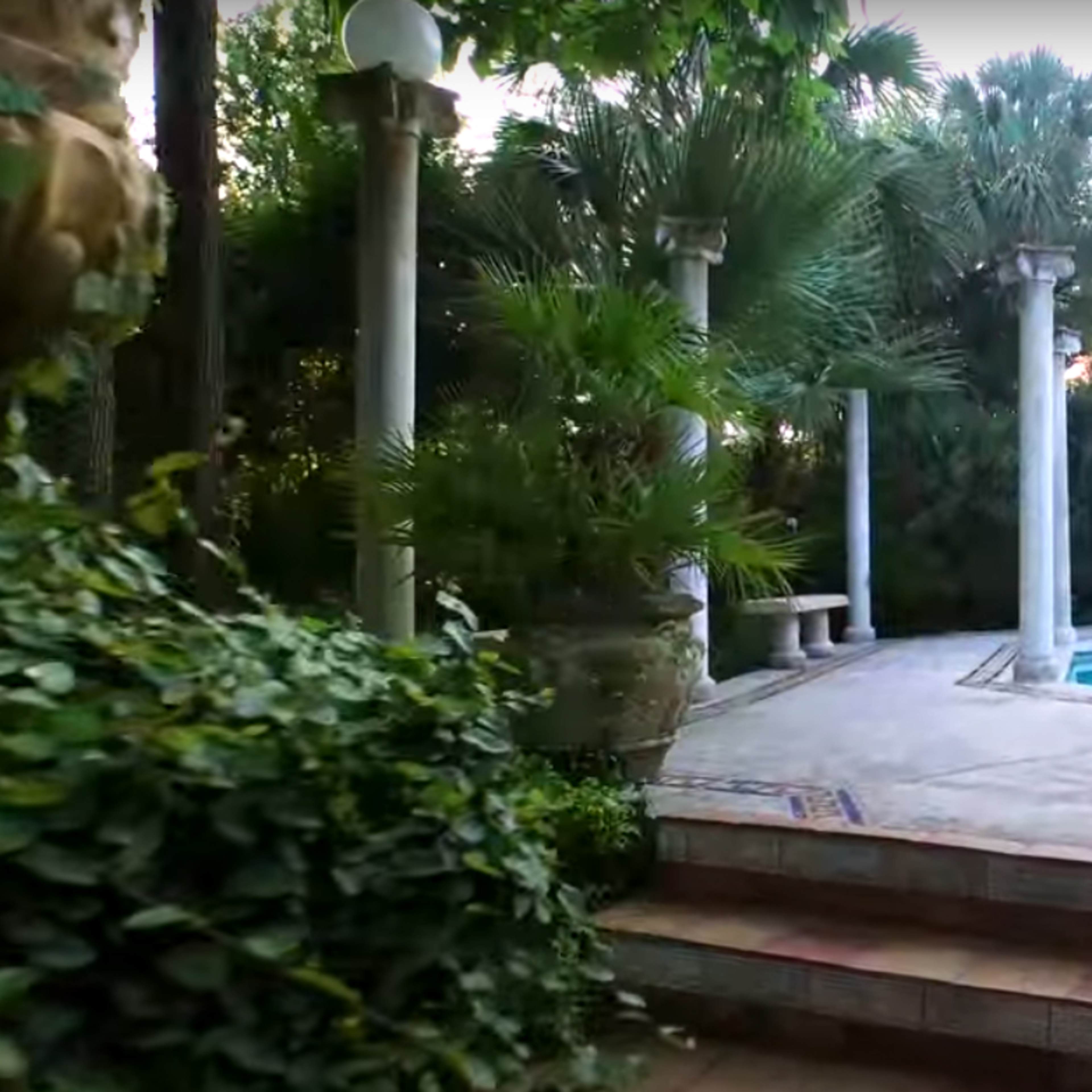 The scene features a tranquil outdoor pool area surrounded by lush greenery and decorative pillars.