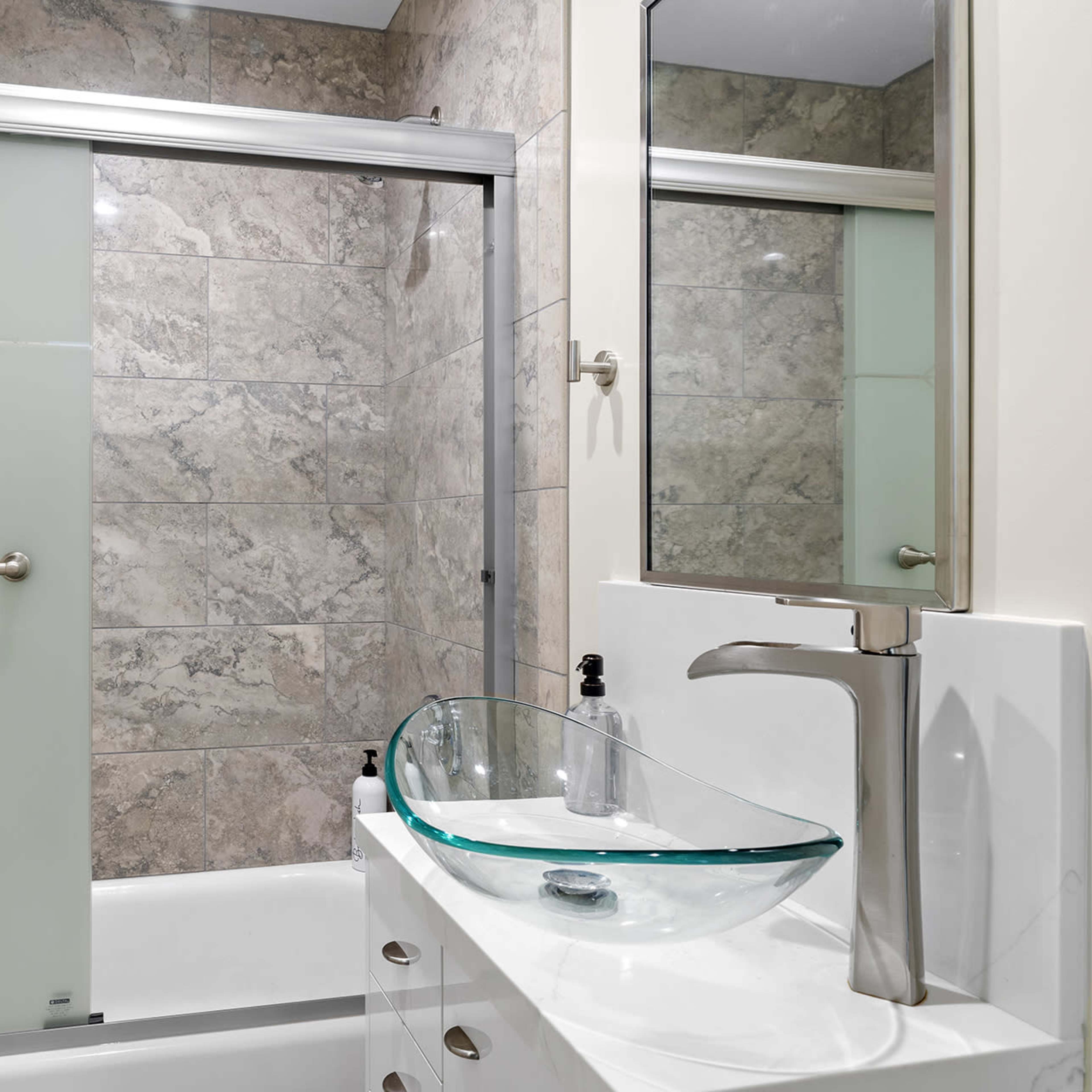 A modern bathroom features a glass shower enclosure, a vessel sink atop a white vanity, and marble-patterned walls.