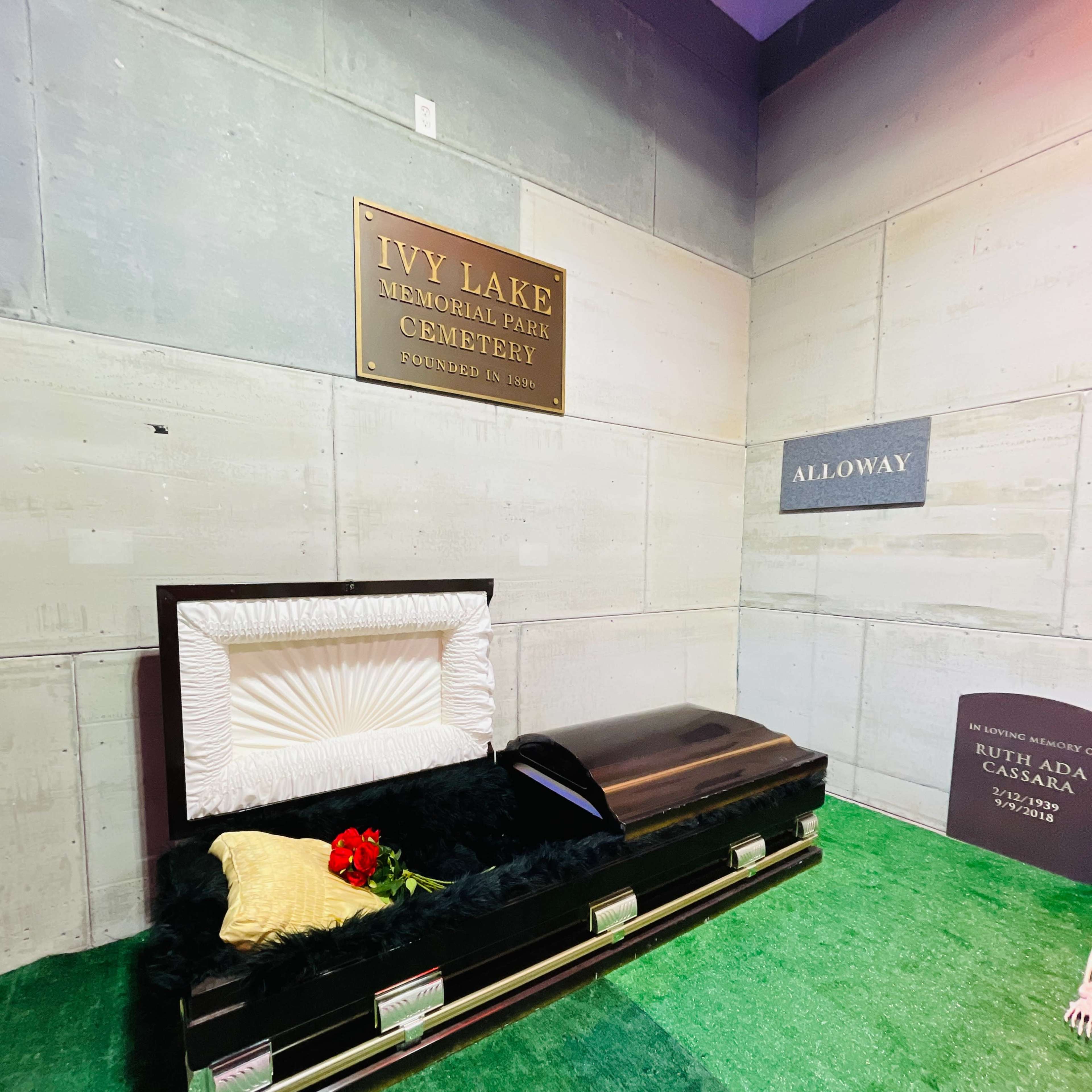An open coffin placed in a cemetery display area, adorned with a red flower and surrounded by a green carpet.