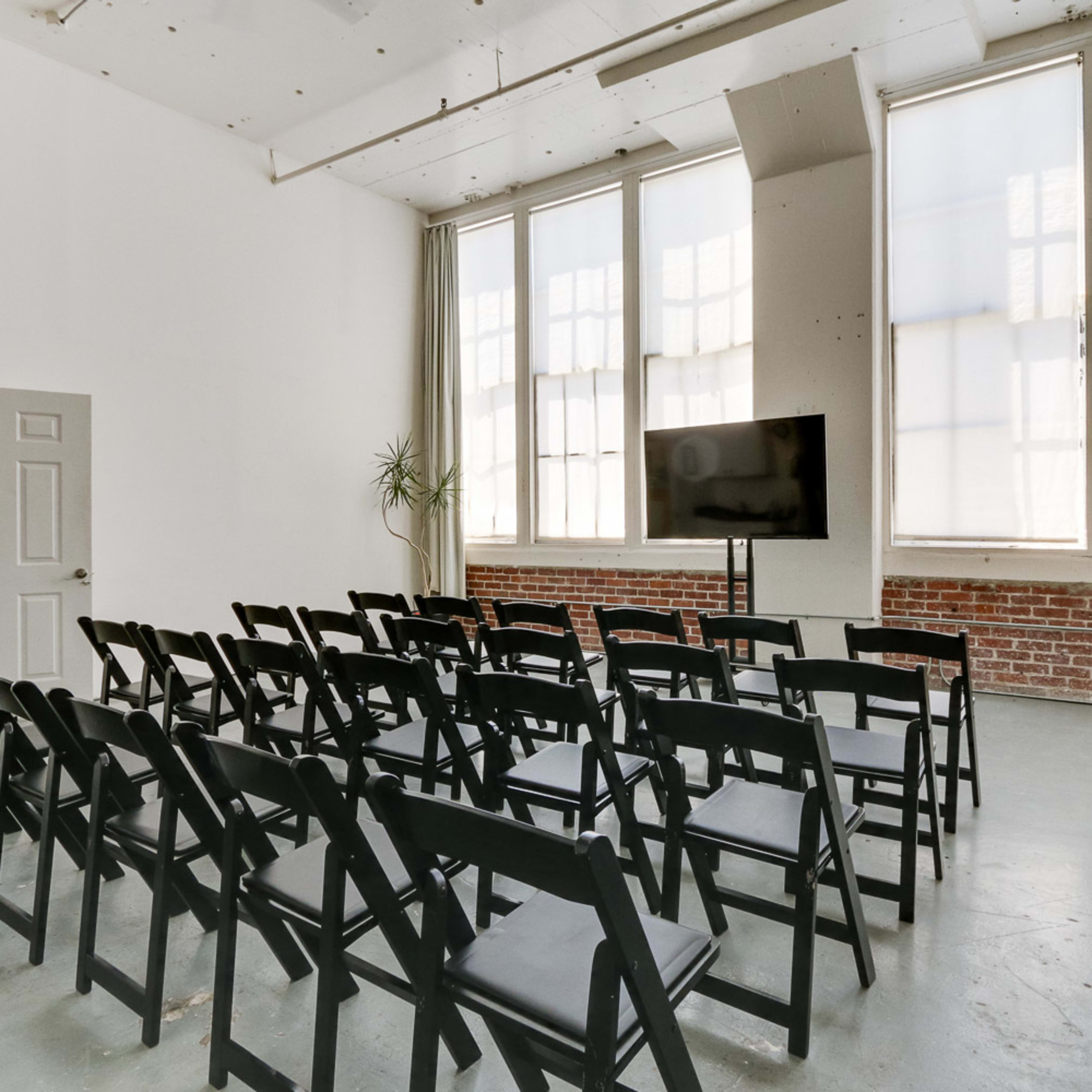 A spacious room with large windows, featuring rows of black folding chairs facing a television on a stand.
