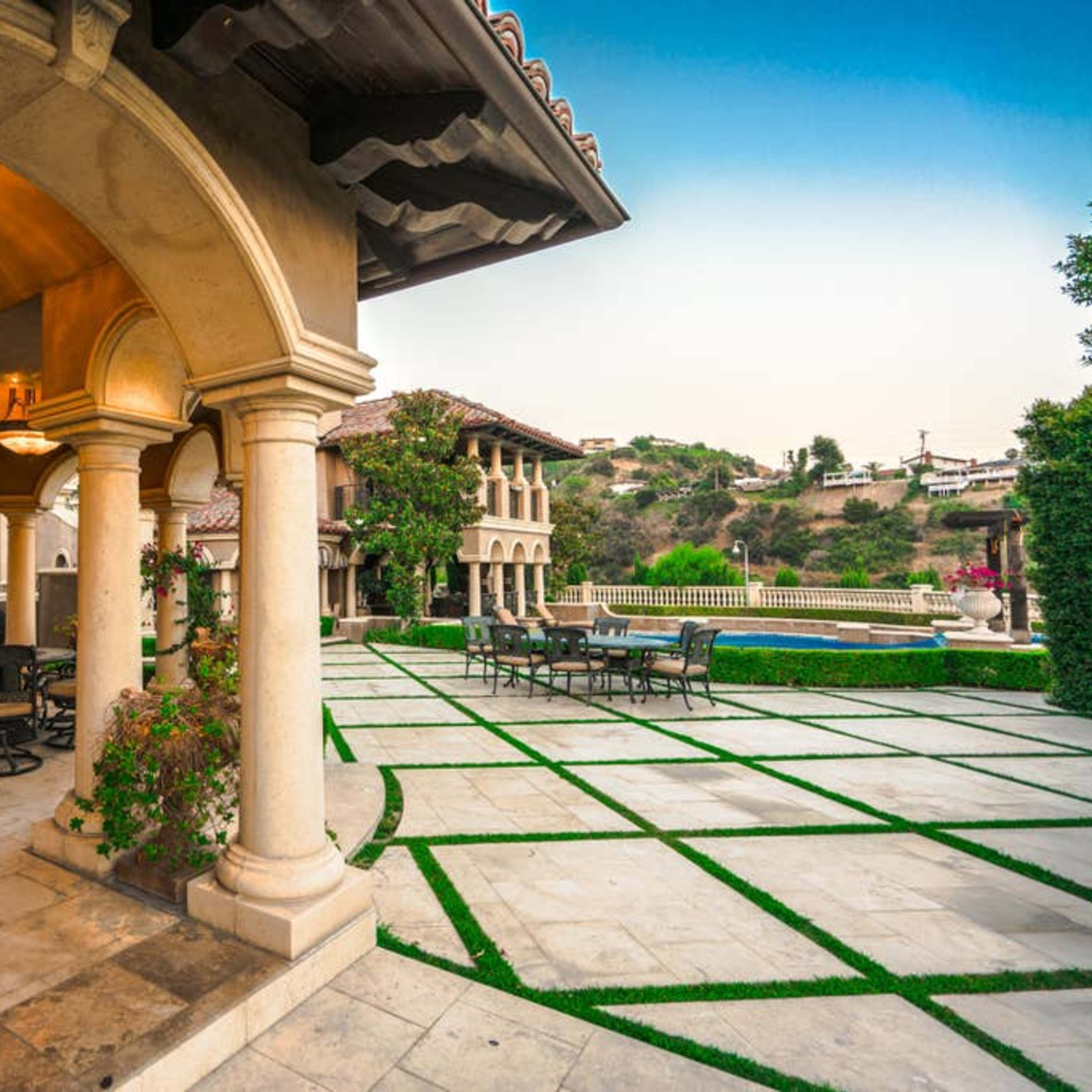 A spacious outdoor area of a luxurious estate, featuring stone columns, outdoor seating, and a landscaped garden with a pool in the background.
