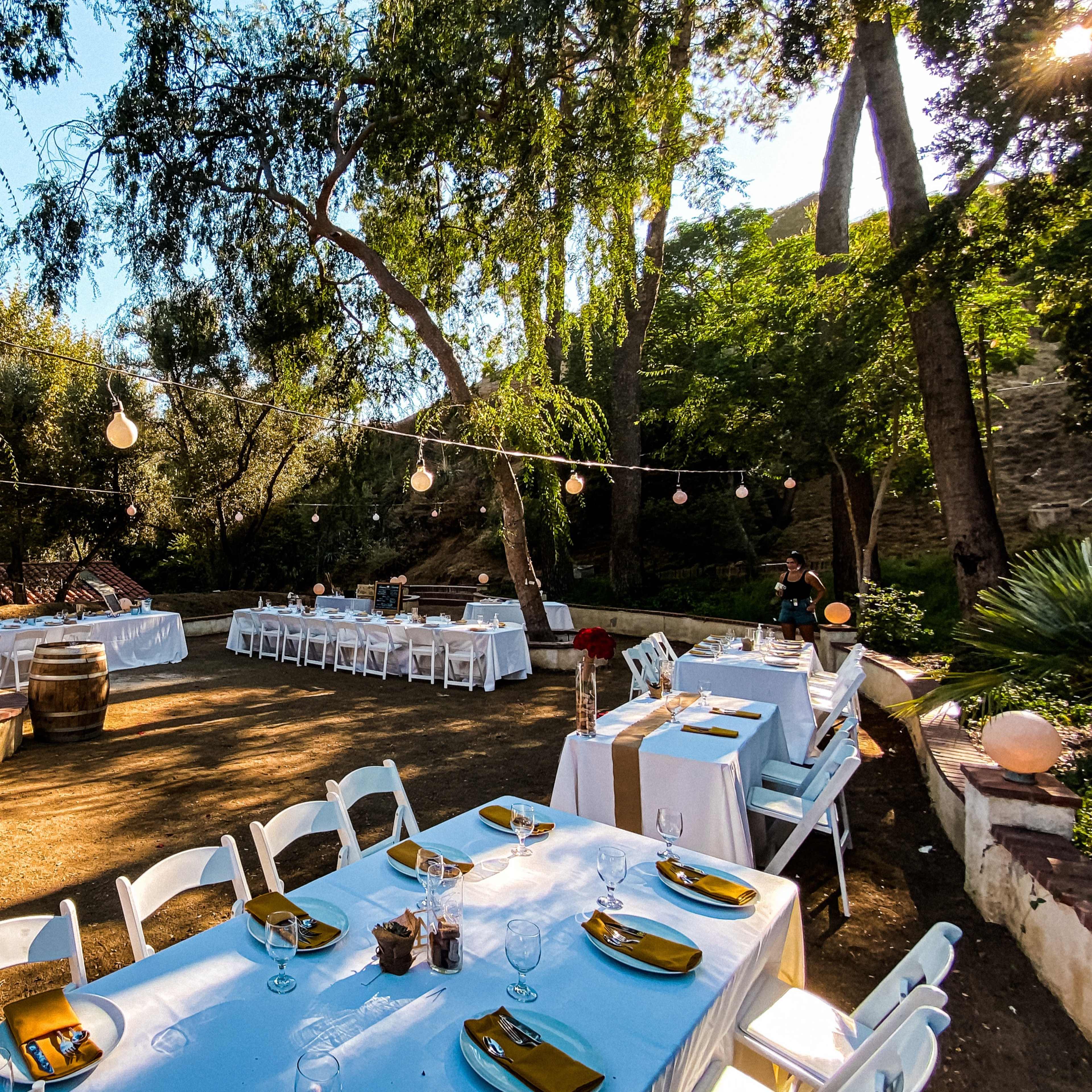 A landscaped outdoor venue set with white tables and chairs under string lights, surrounded by trees.