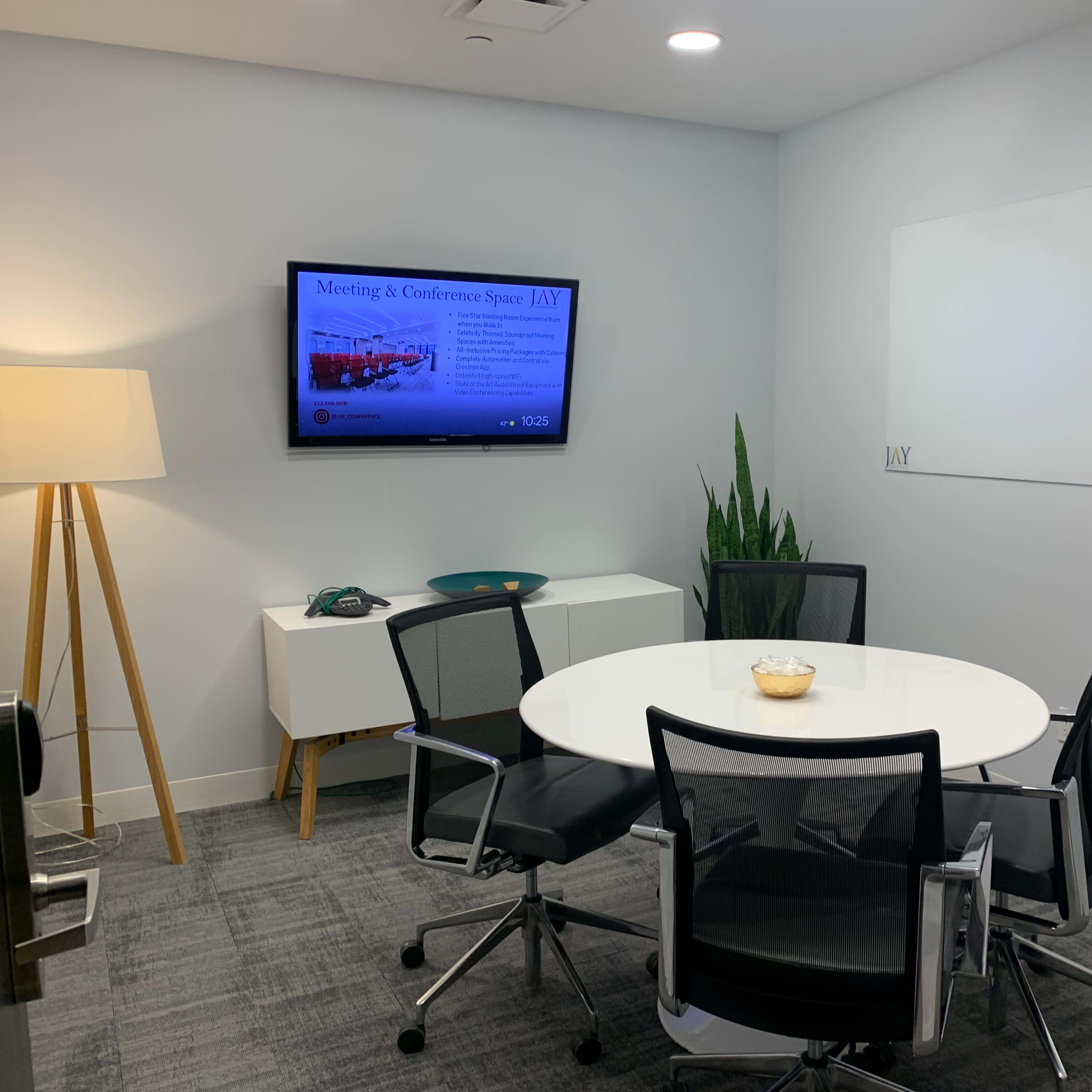 A modern conference room features a round table with four chairs, a television displaying meeting information, and a potted plant in the corner.