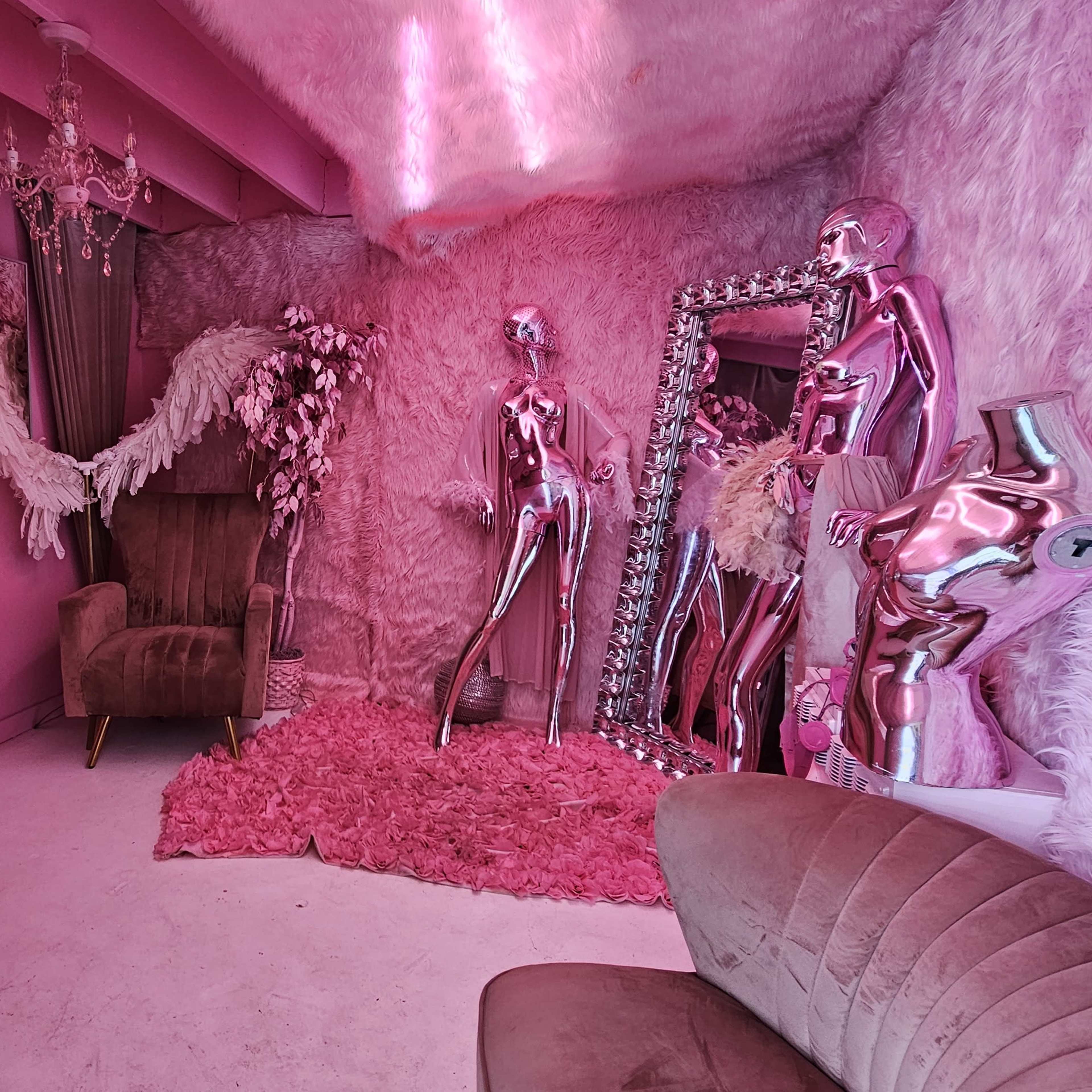 A vibrant room features pink walls, a plush rug, metallic mannequins, a mirrored wall, and elegant furniture.