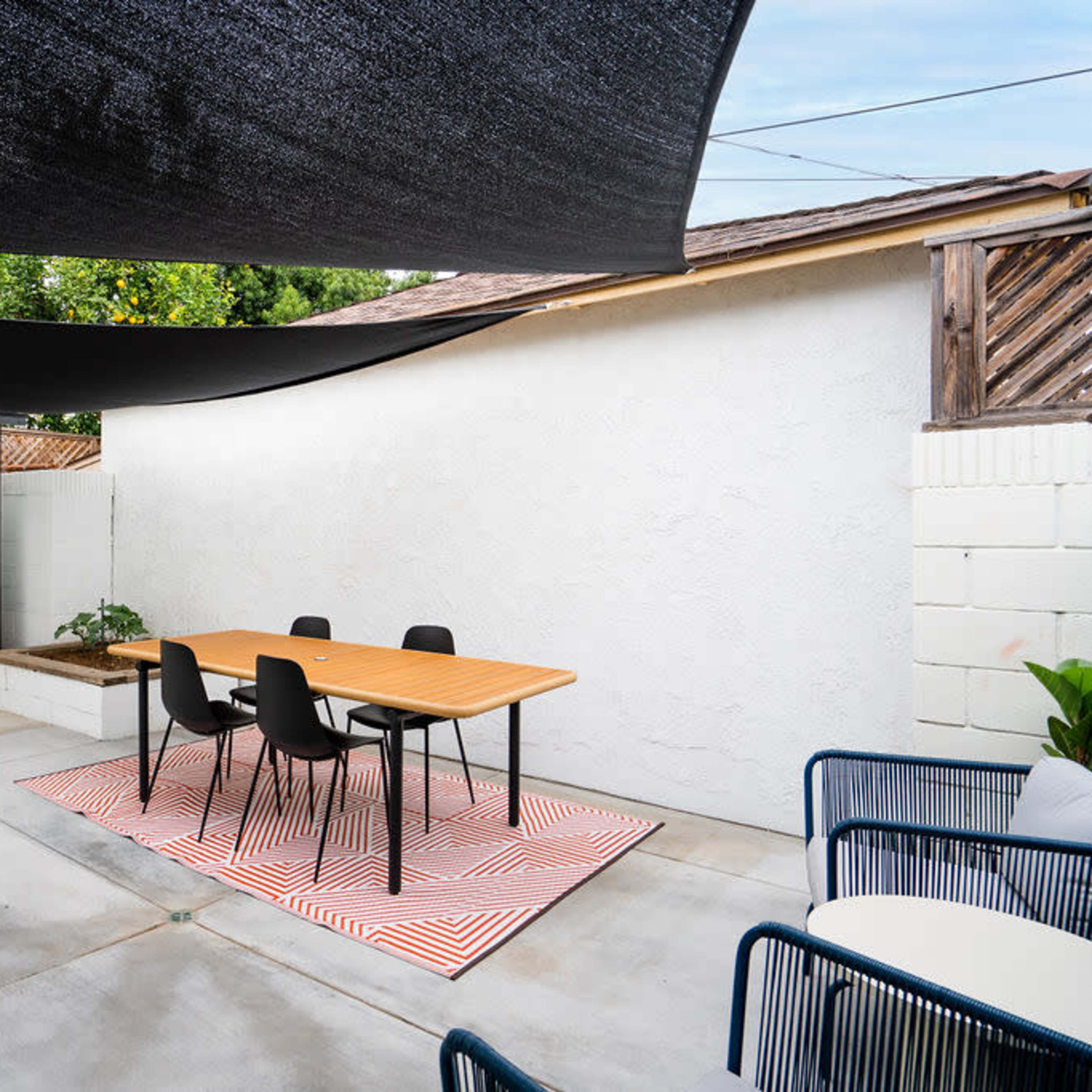 A minimalist outdoor dining area with a wooden table and black chairs, sheltered by black shade sails, adjacent to a white wall.