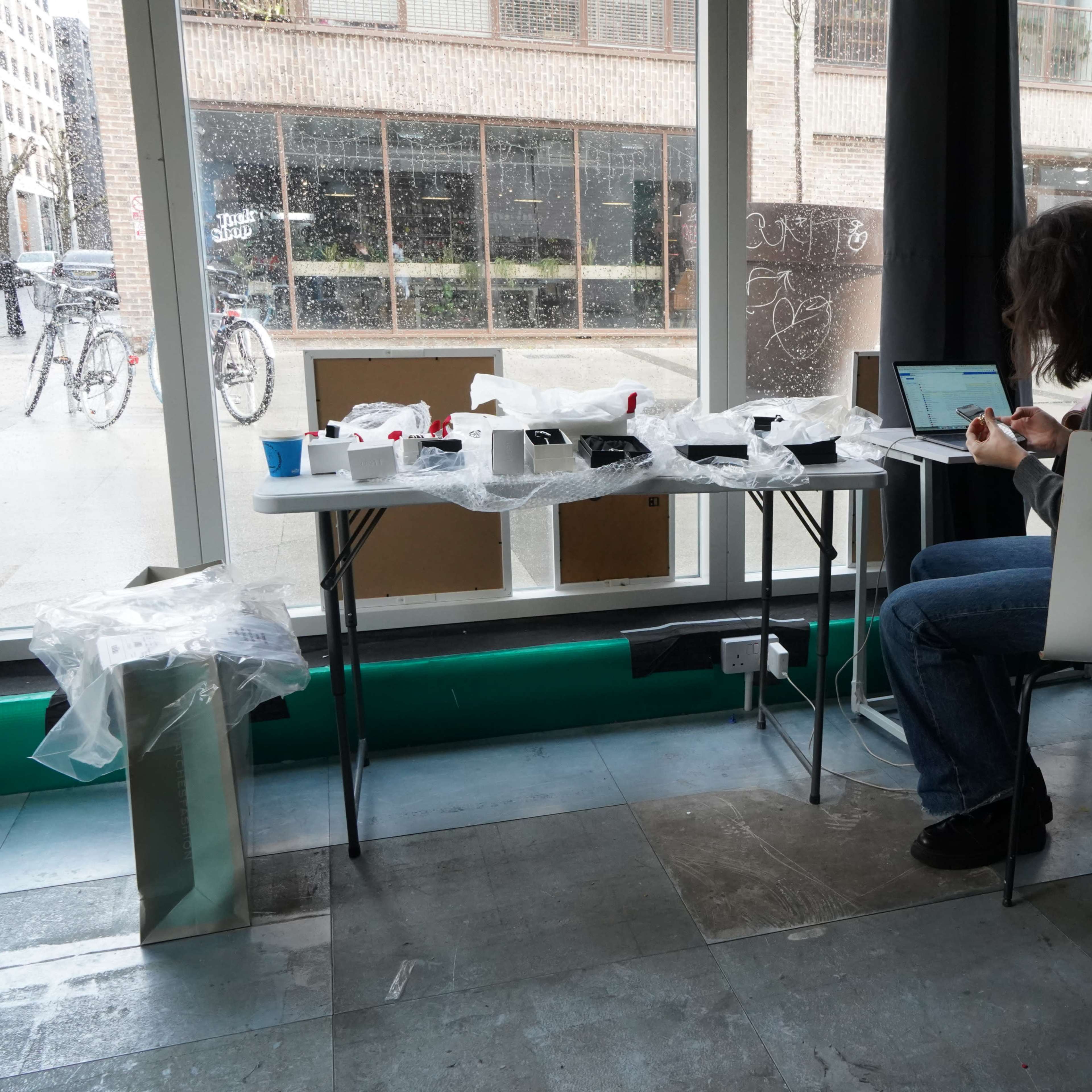 A person works at a table covered in plastic wrap next to a large window, with various objects and a laptop in front of them.