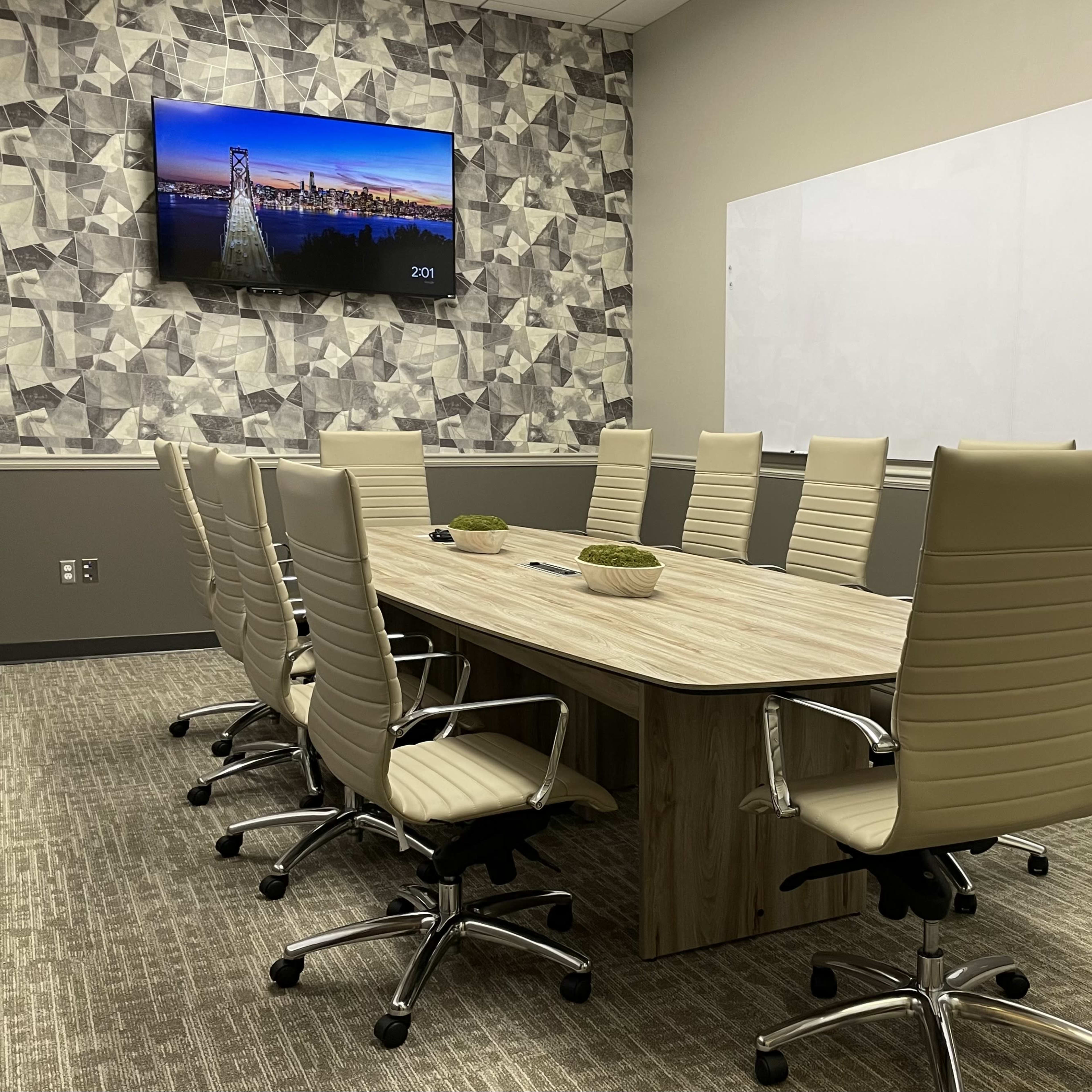 A modern conference room features a long wooden table surrounded by eight light-colored ergonomic chairs, with a television mounted on the wall displaying a cityscape.