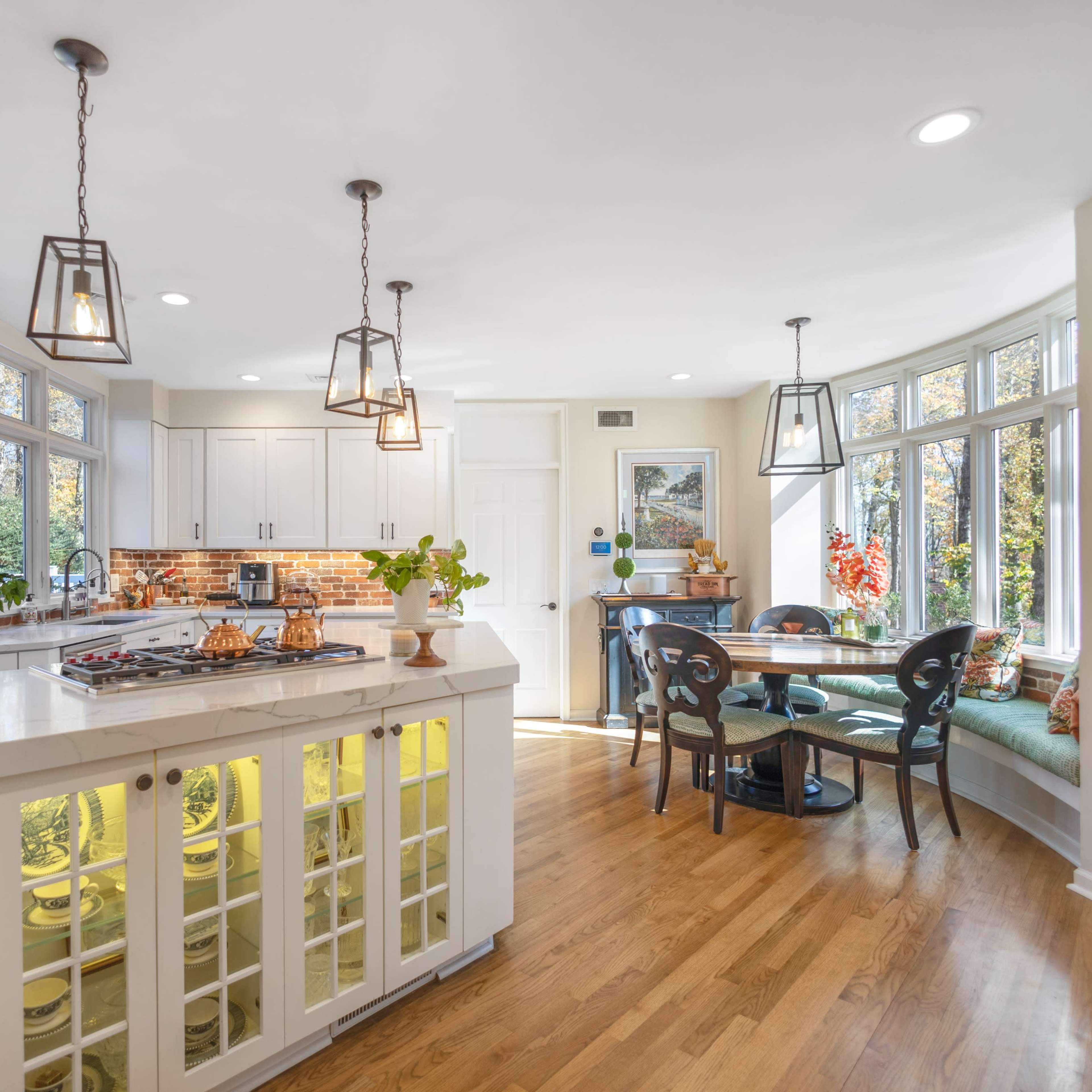 A bright kitchen with a central island featuring a stovetop, surrounded by large windows and a cozy breakfast nook.