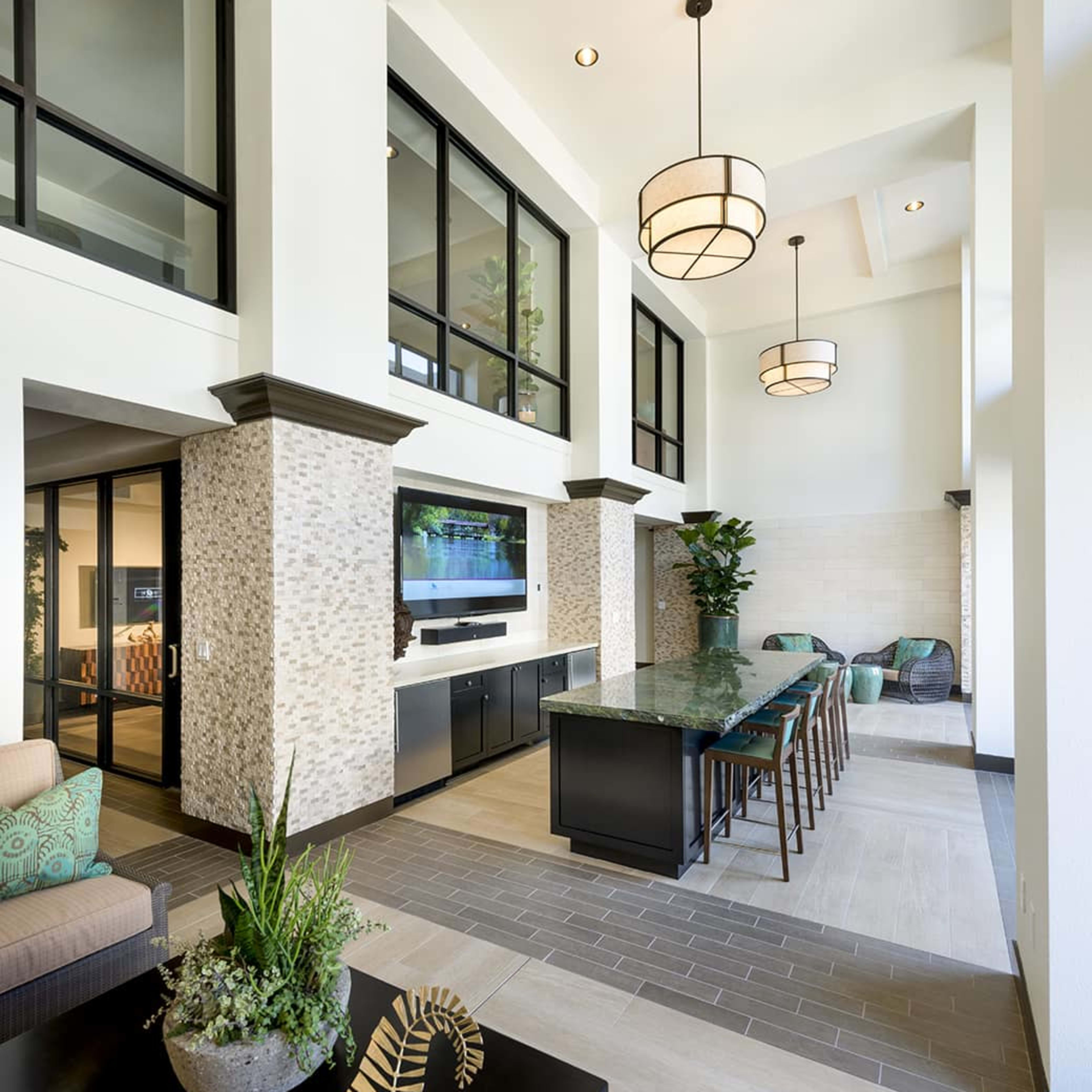 A modern communal area with a high ceiling, featuring a long table, contemporary lighting fixtures, and large windows.