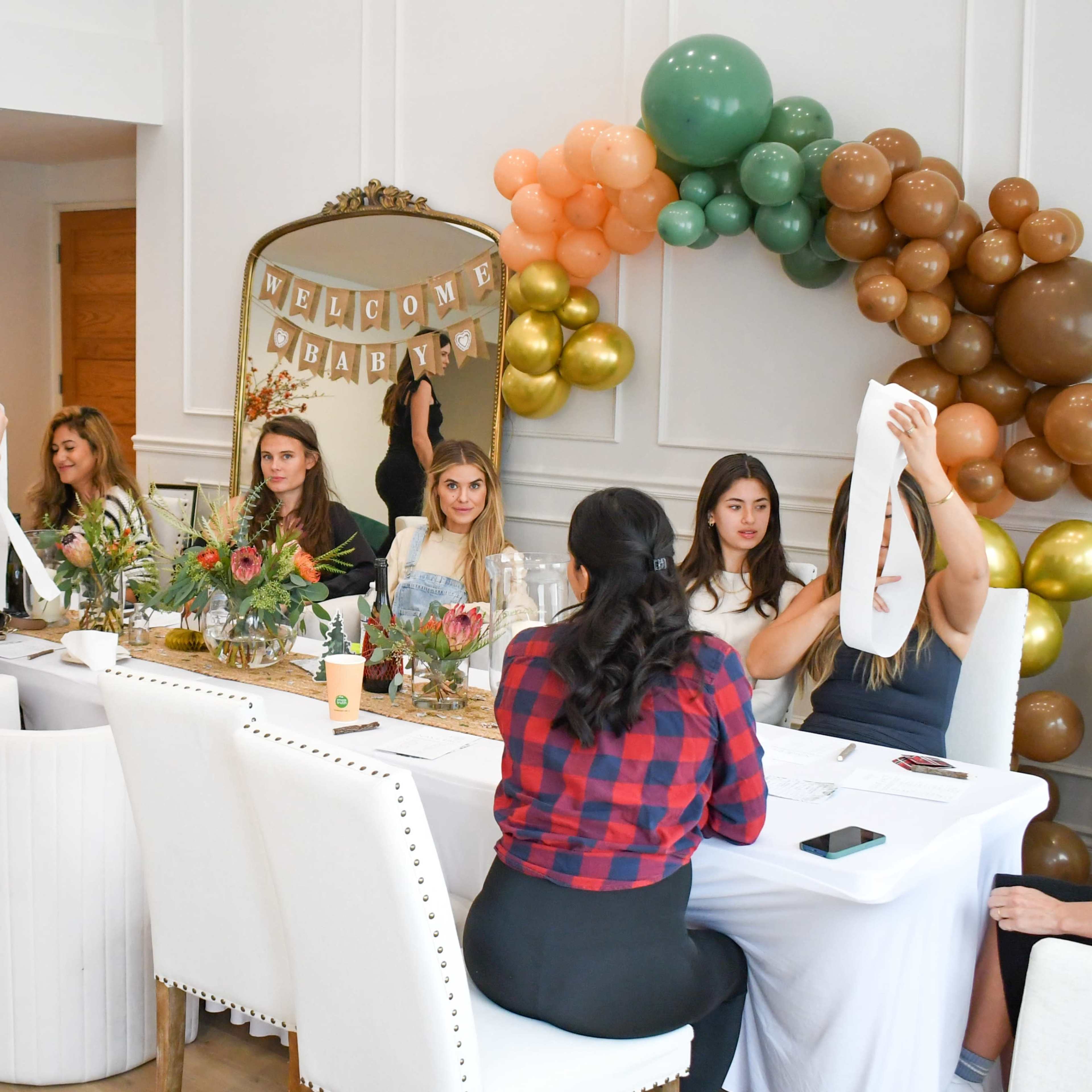 A group of women sits around a table decorated with floral arrangements and balloons, holding up cloth items while a woman stands in the background.