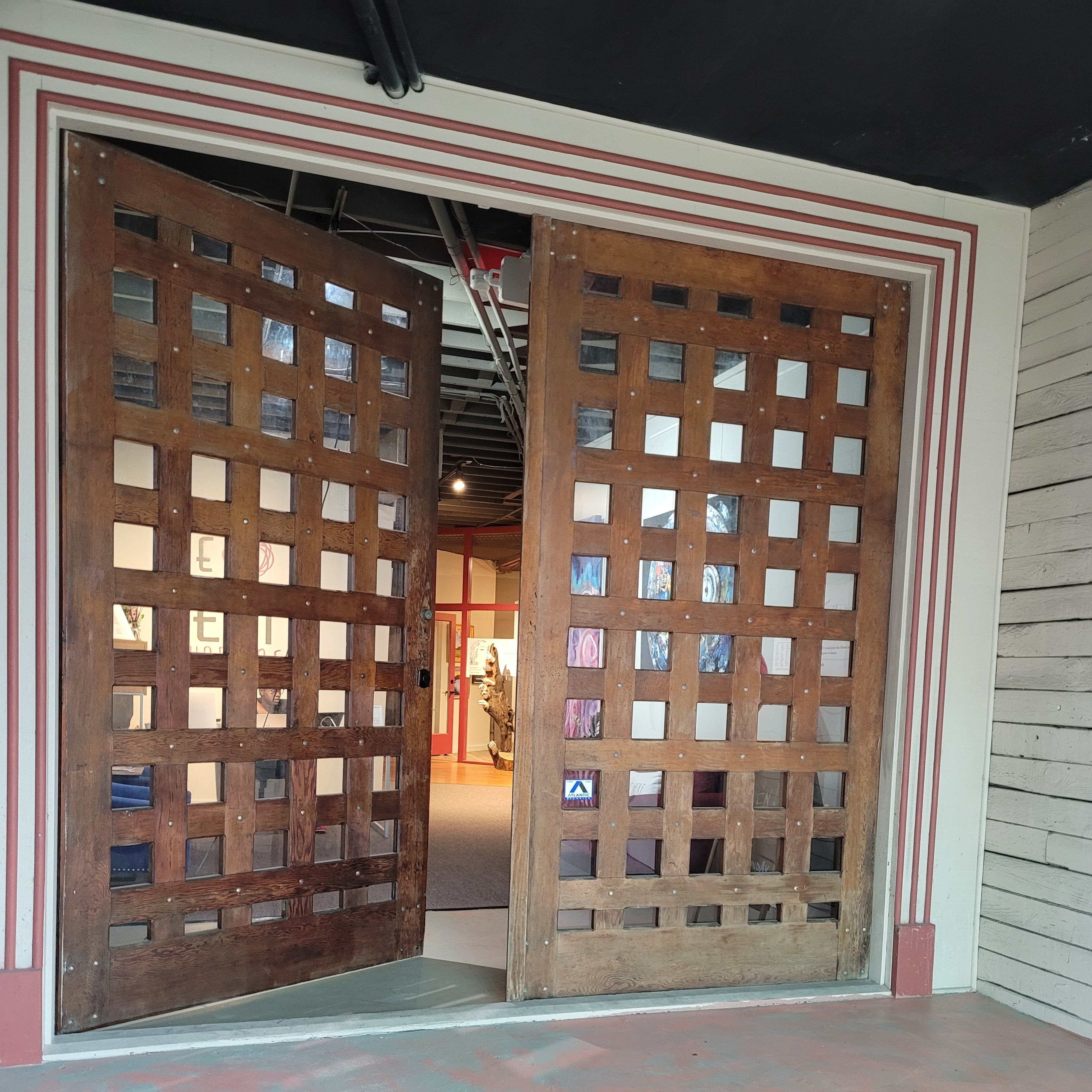 A set of large, wooden double doors with a grid pattern, partially opened to reveal a hallway and gallery beyond.