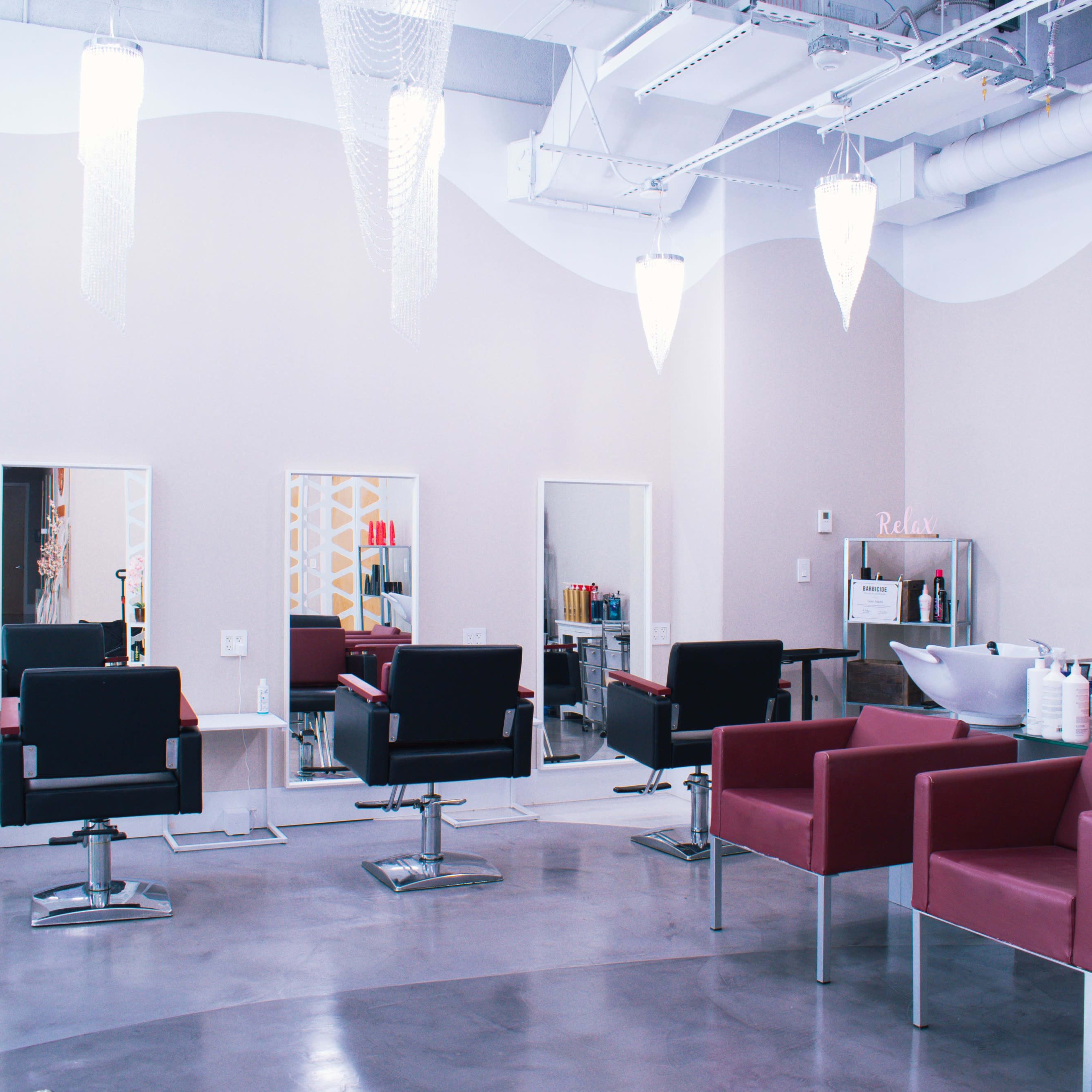 A modern salon features several black styling chairs, white mirrors, and two shampoo bowls in a spacious, well-lit interior.