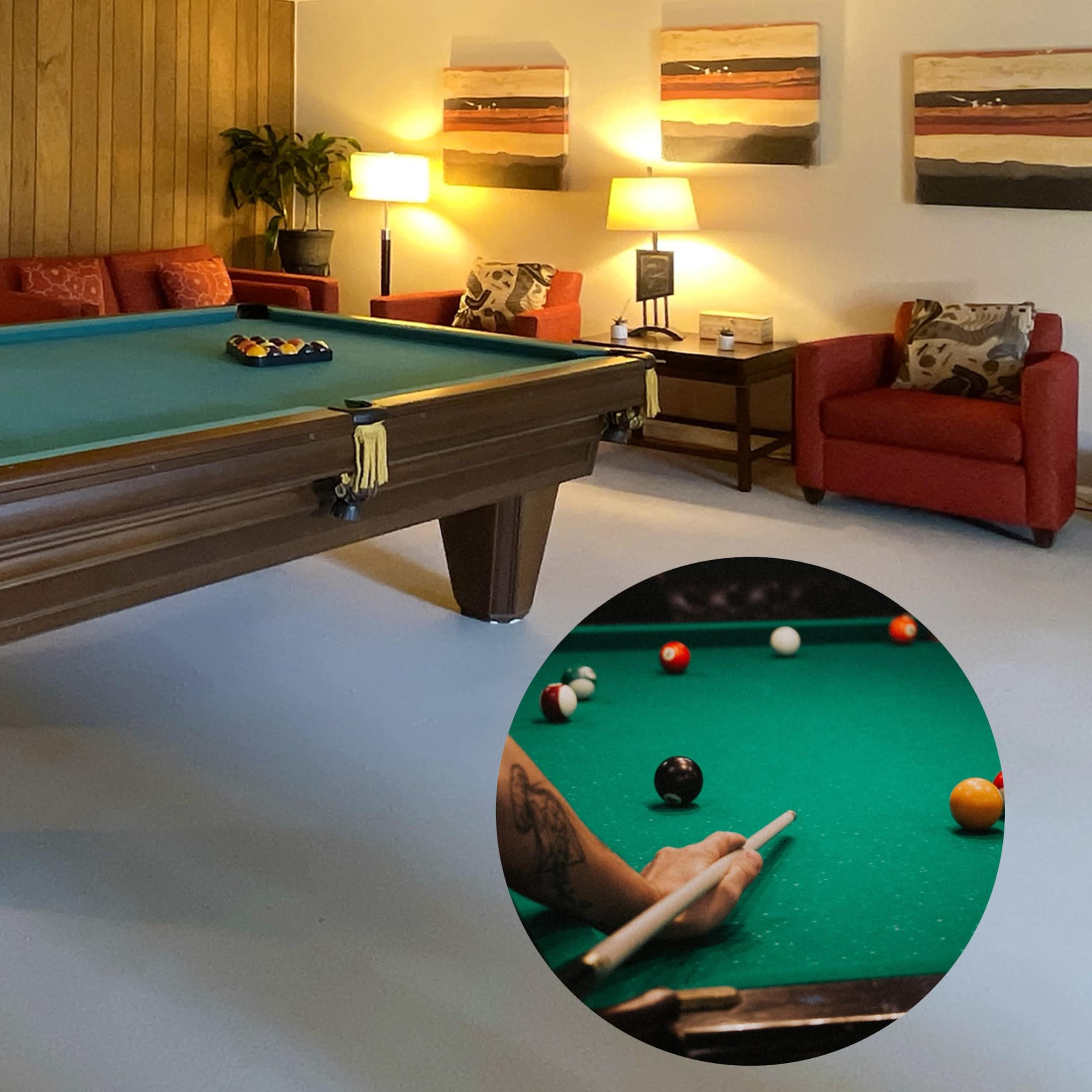 A game room featuring a green billiards table, two red armchairs, a mirror on the wall, and several framed artworks.