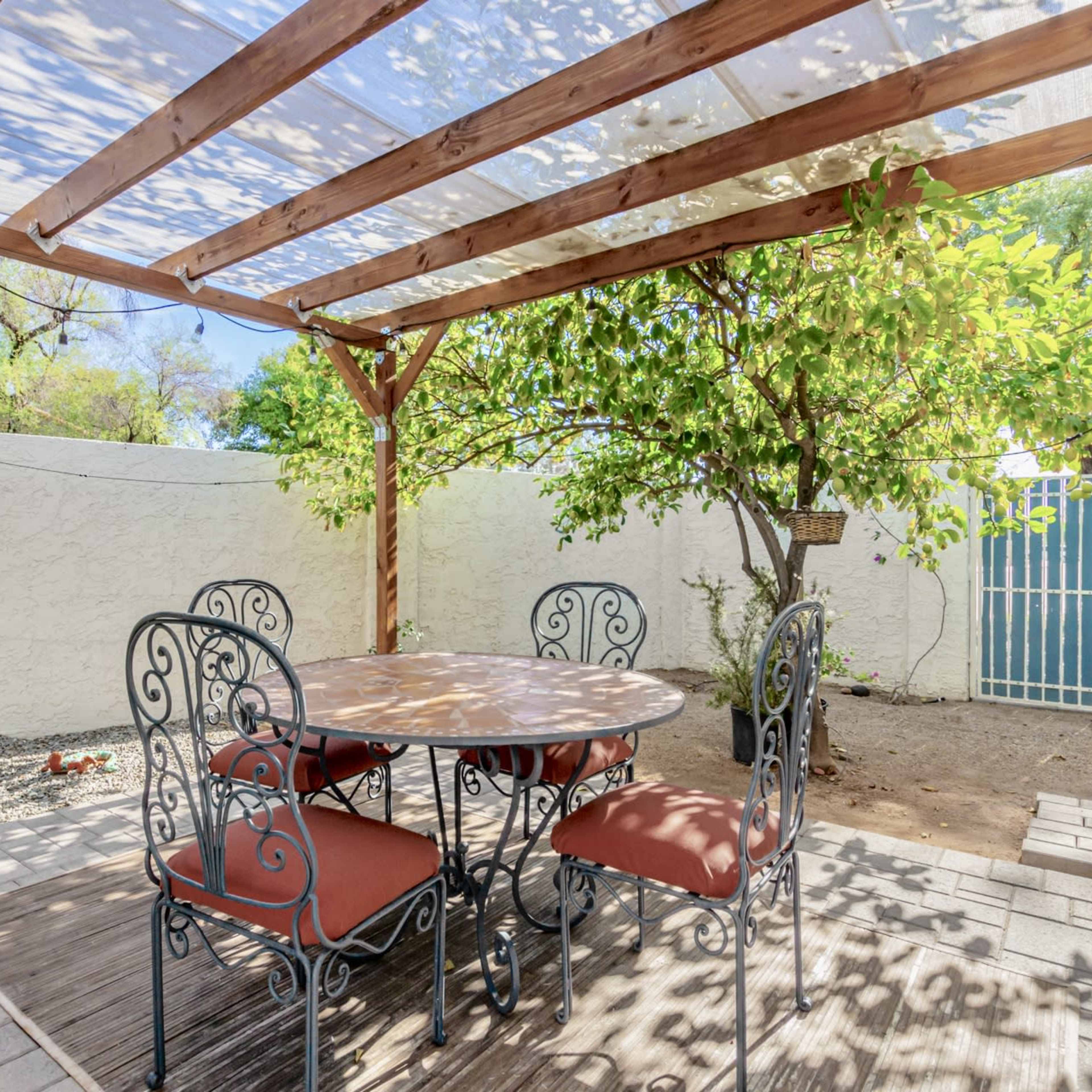 A covered outdoor dining area with a round table and four chairs, surrounded by green trees and a fenced yard.