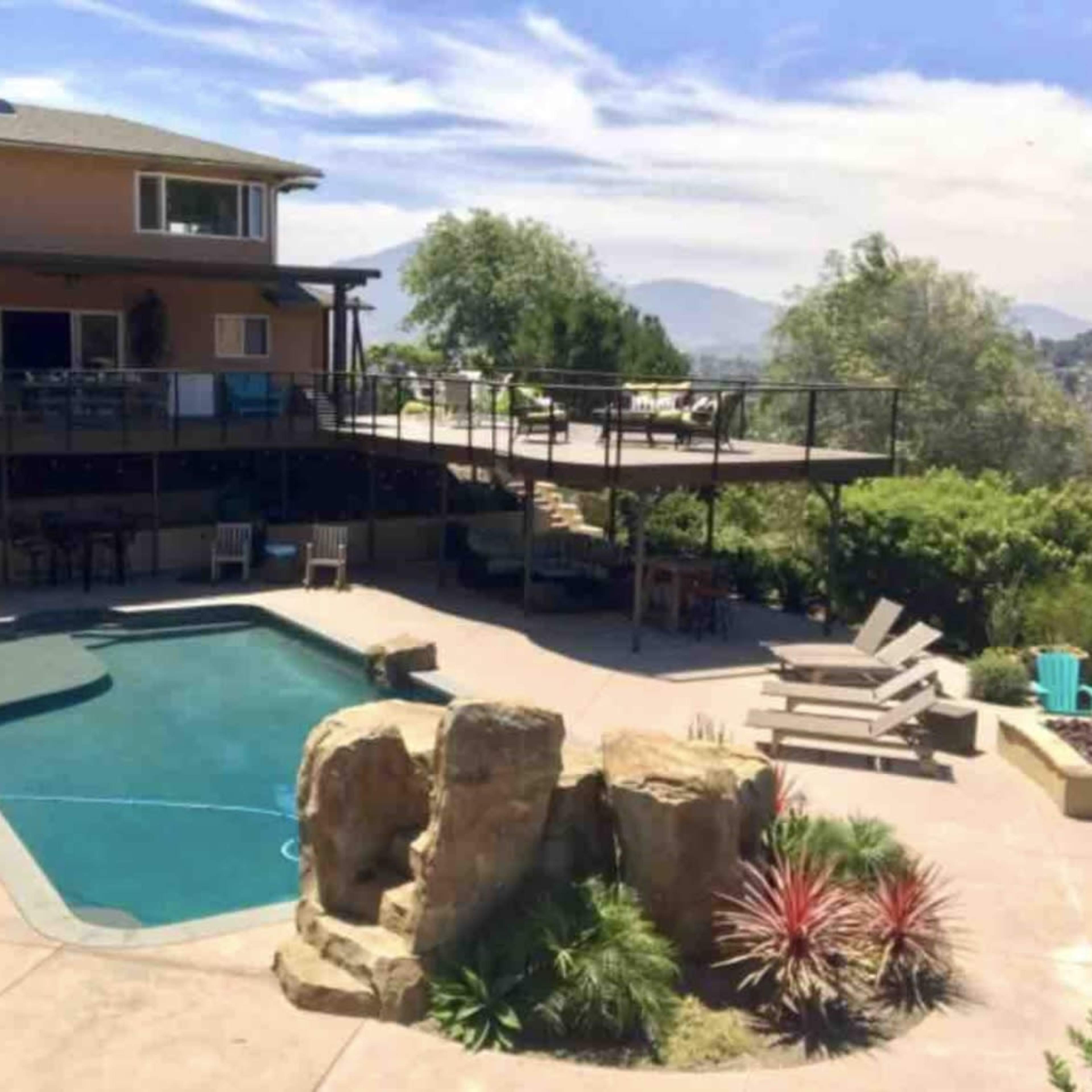 A house with a deck overlooks a swimming pool and landscaped yard featuring rocks and plants.