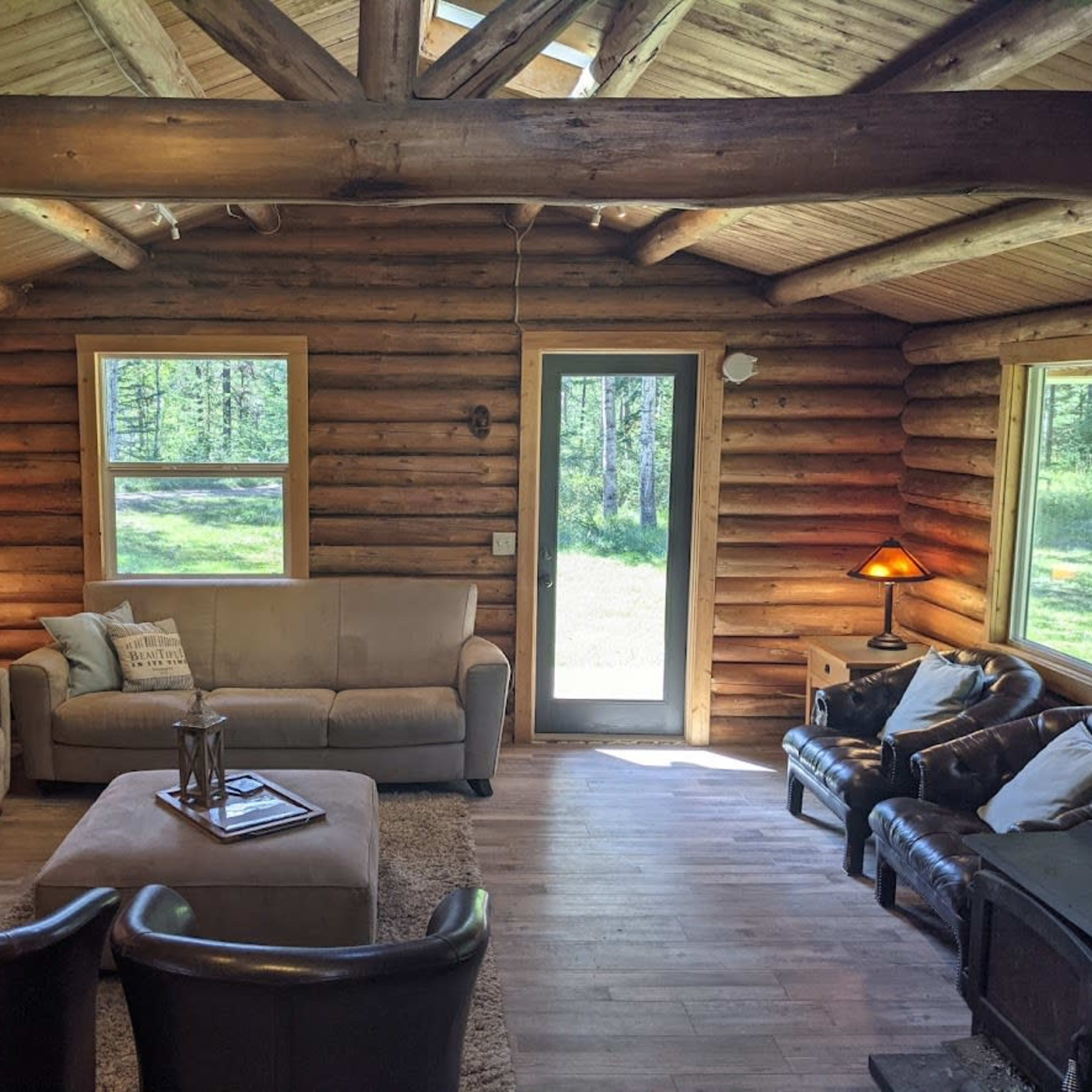 The interior of a log cabin living room features a beige sectional sofa, a circular coffee table, two dark armchairs, and large windows providing a view of the outdoor greenery.
