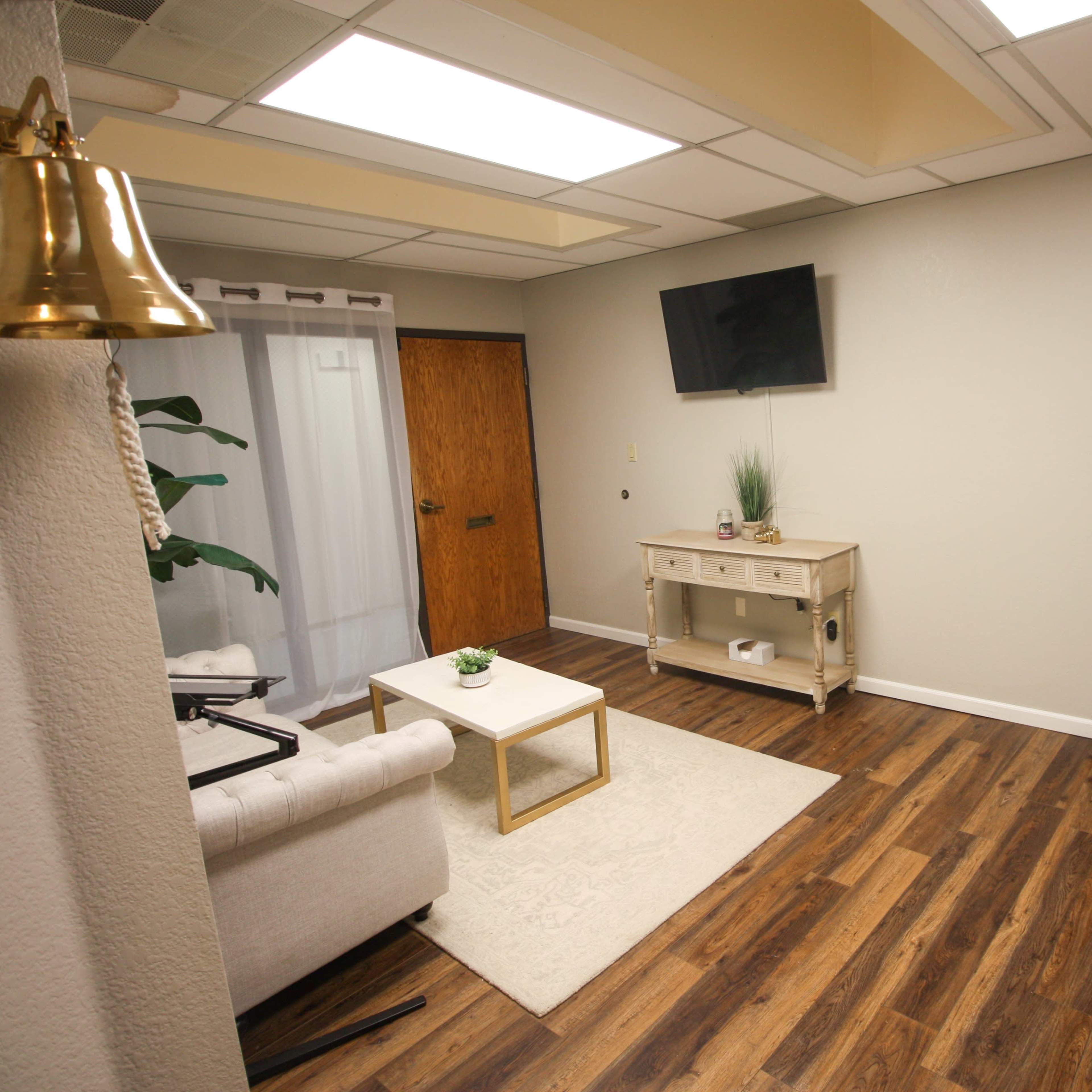A well-lit room featuring a sofa, a coffee table, a television on the wall, and wooden flooring.
