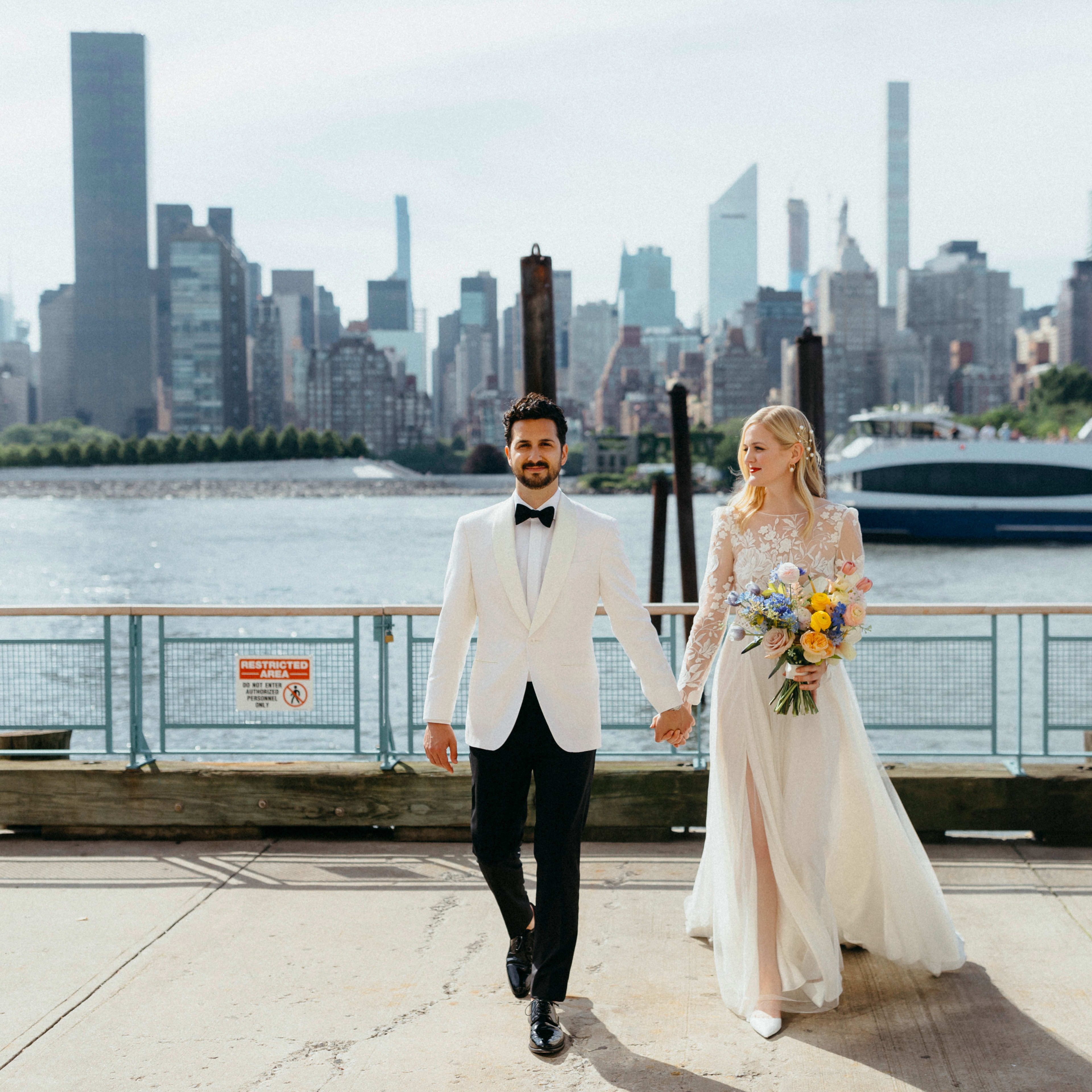 A bride and groom walk hand in hand along a waterfront promenade with a skyline view of New York City in the background.