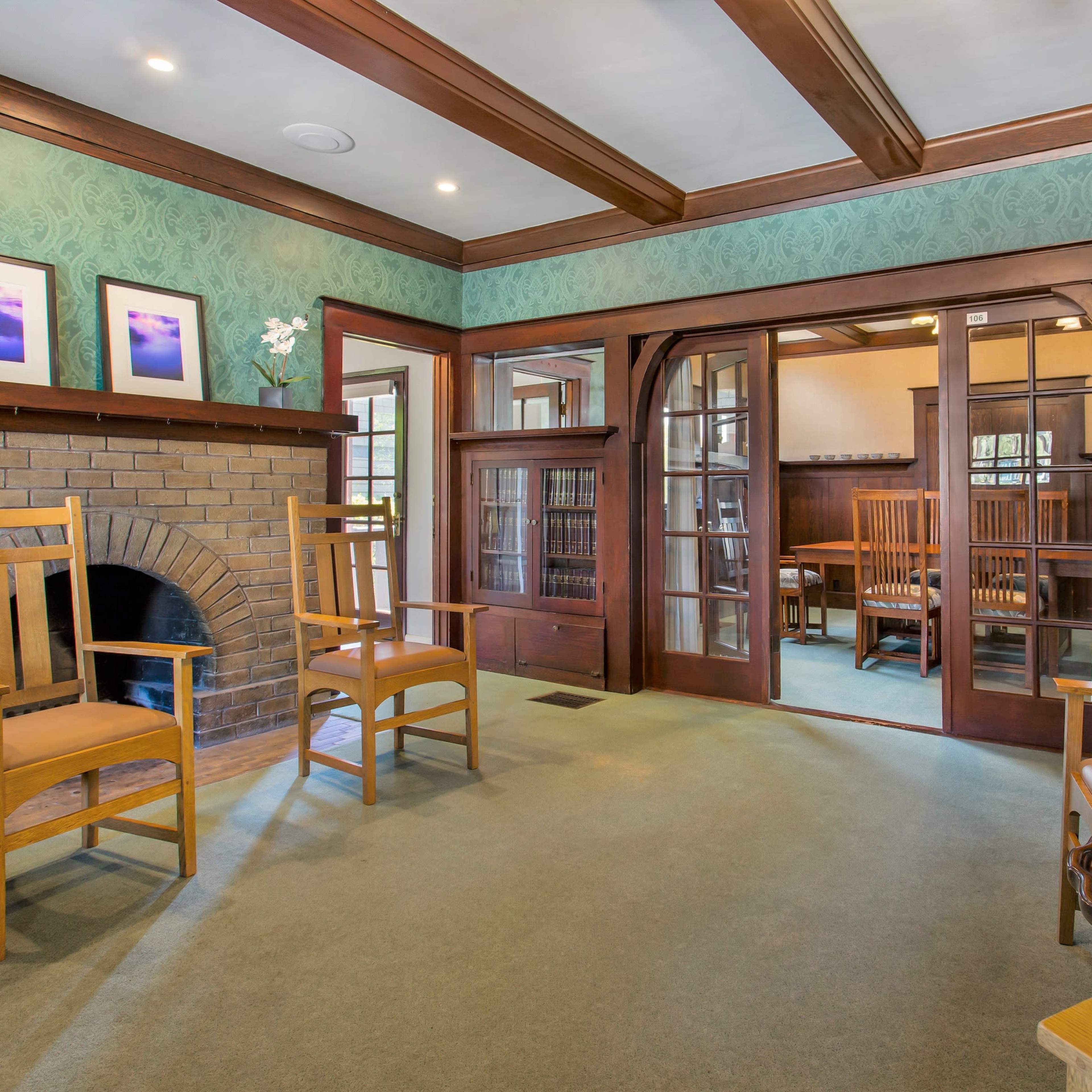 A room with wooden beams on the ceiling, two wooden chairs placed in front of a fireplace, and large glass doors leading to another seating area lined with bookshelves.