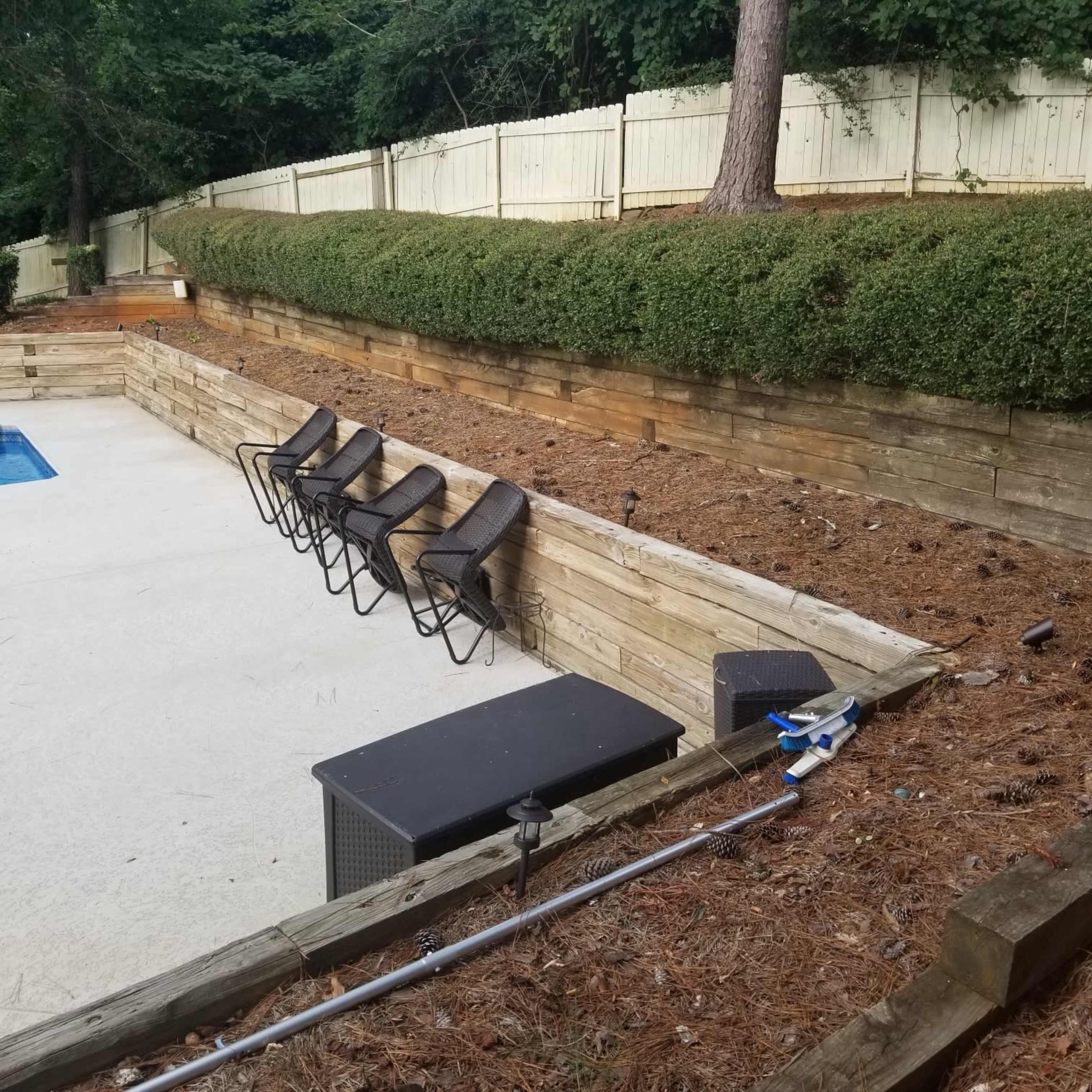 A pool area with four lounge chairs arranged beside a partially filled pool surrounded by a wooden retaining wall and a carpet of pine needles.