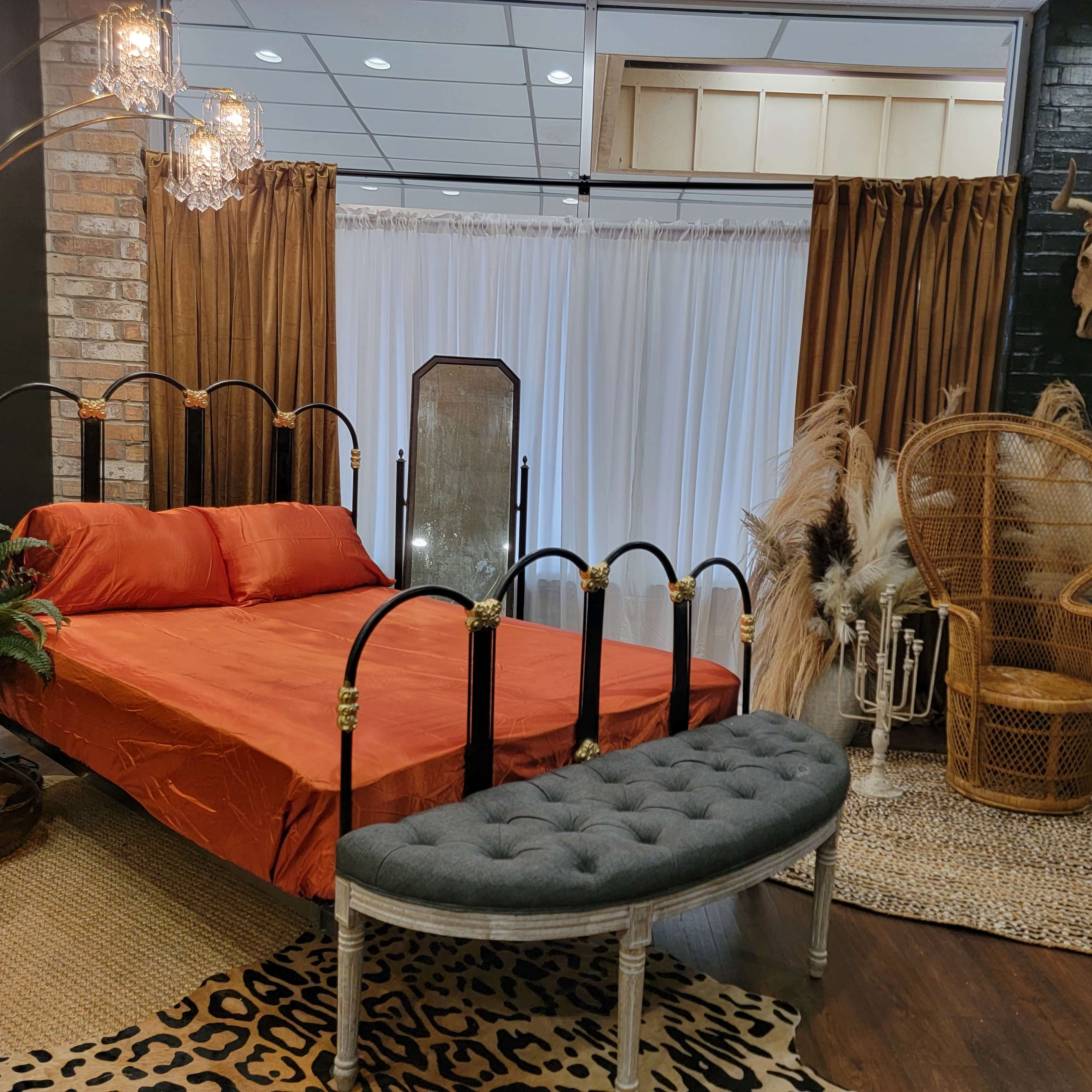 A modern bedroom setup features an orange bed with a metal frame, a decorative mirror, and a rattan chair surrounded by plants and patterned rugs.