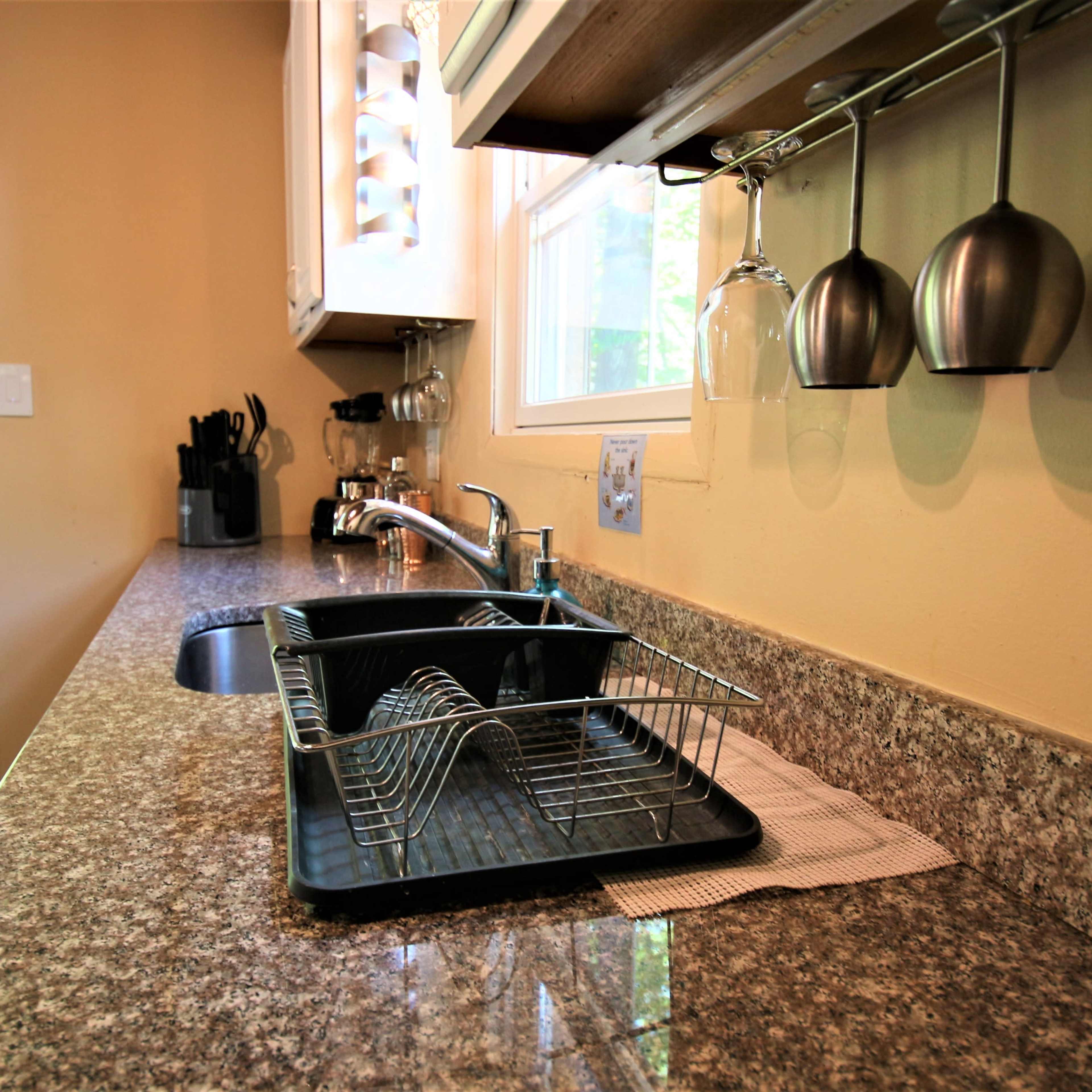 A kitchen countertop features a dish rack with clean dishes, a sink, and hanging stainless steel cups under a cabinet.