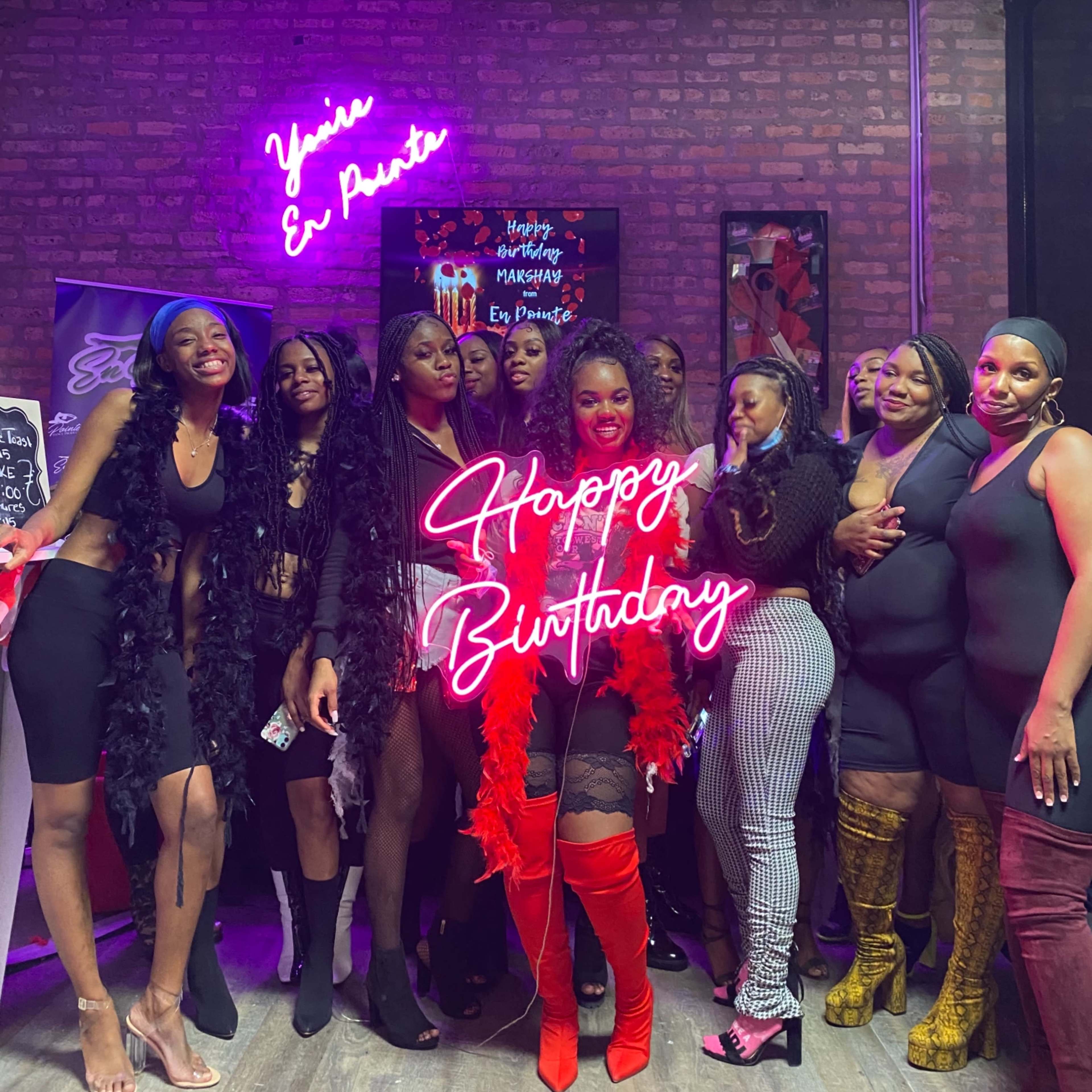 A group of women poses together in a brightly lit venue with a neon "Happy Birthday" sign.