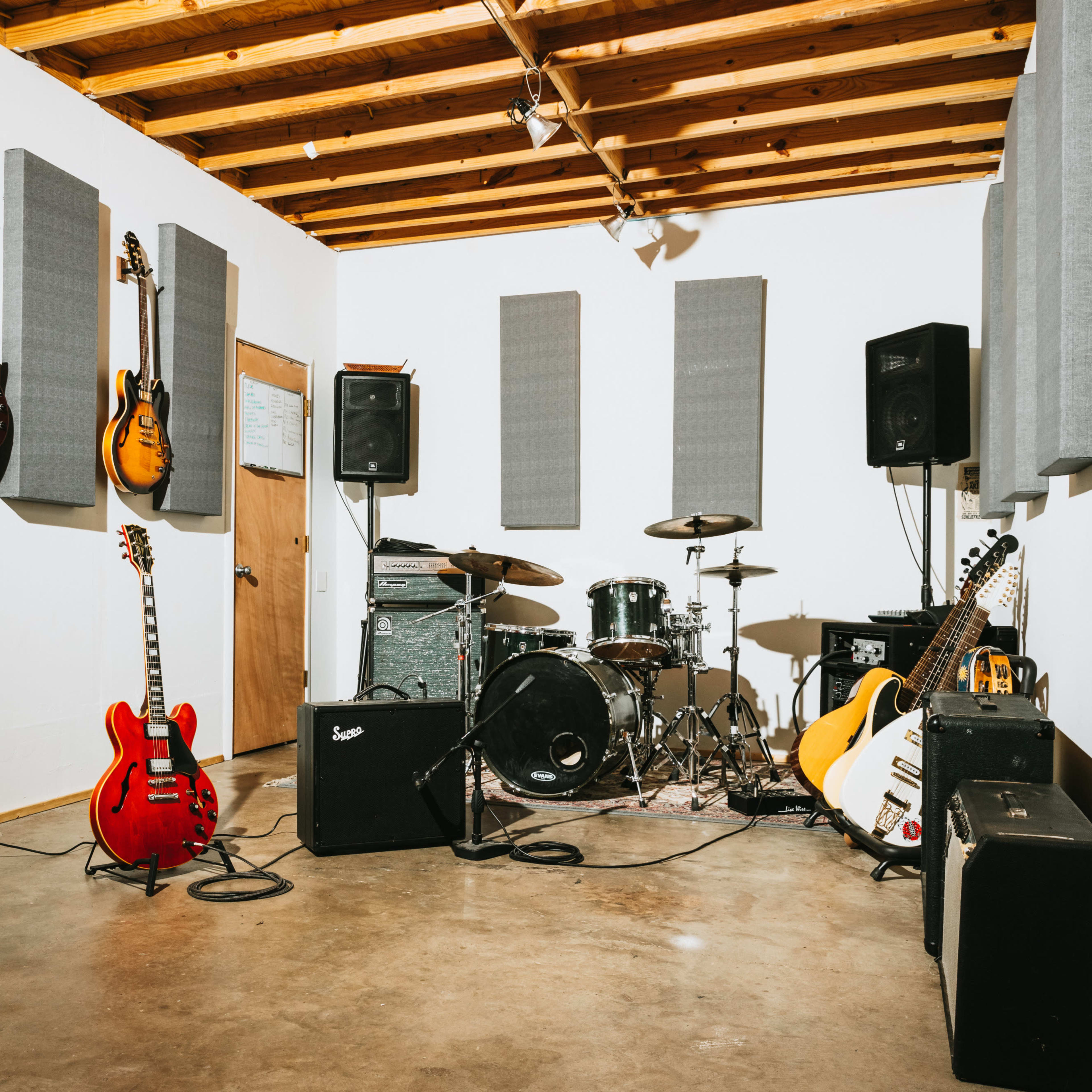 A music rehearsal space featuring guitars on the walls, a drum set, amplifiers, and soundproofing panels.