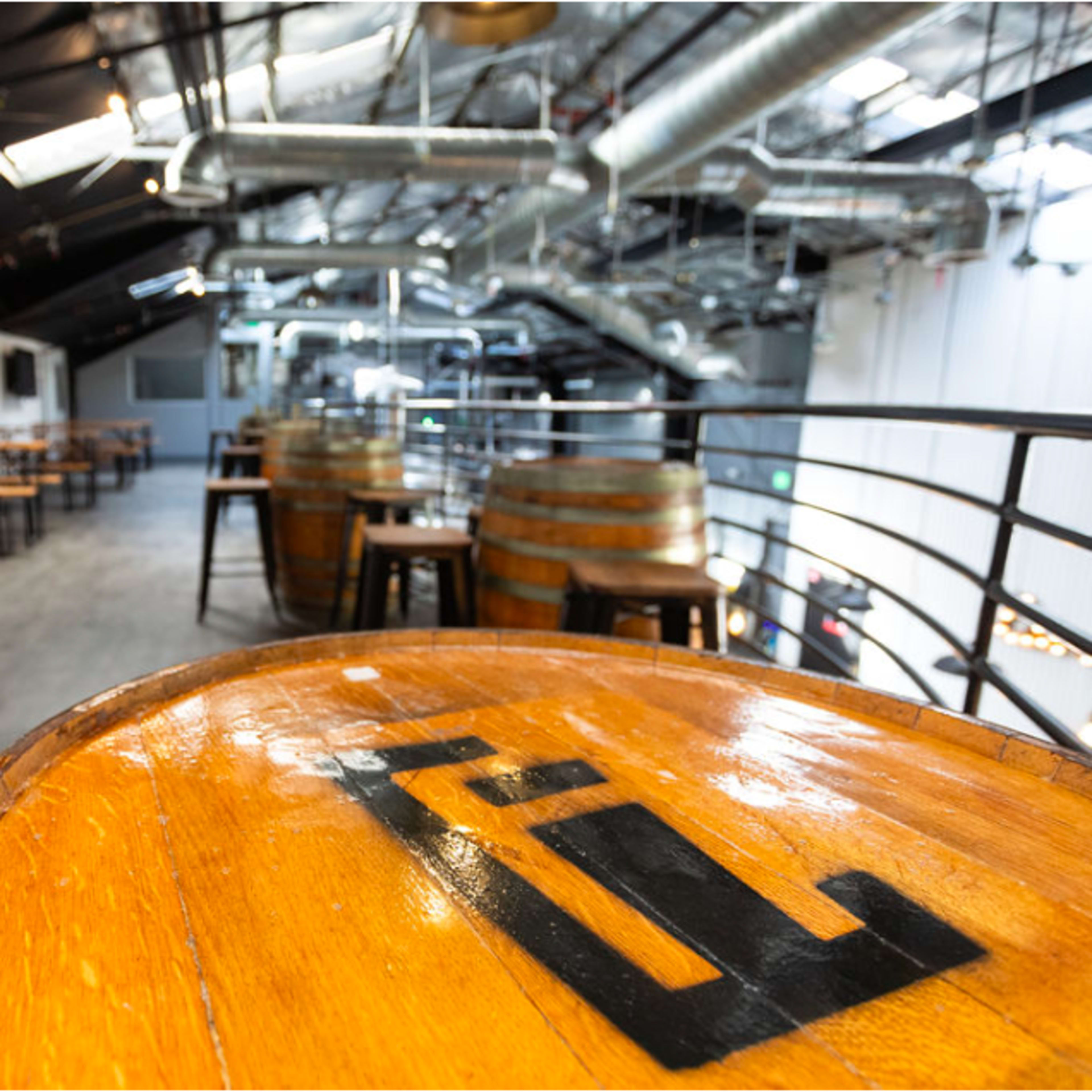 An interior of a bar with wooden tables and barrels, featuring exposed metal ductwork and a TV on the wall.