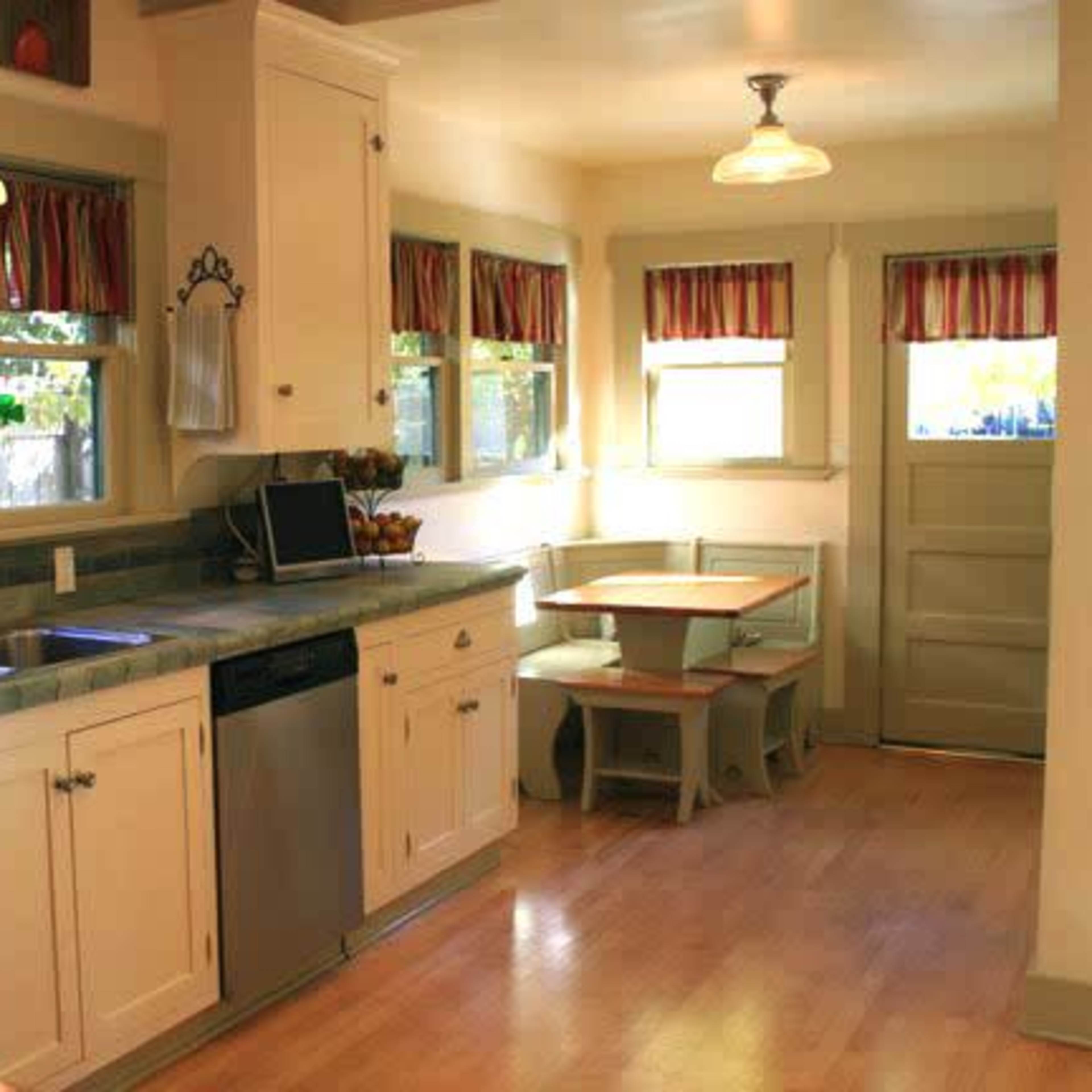 A kitchen features a tiled countertop, white cabinets, and a small dining table beside a set of windows.