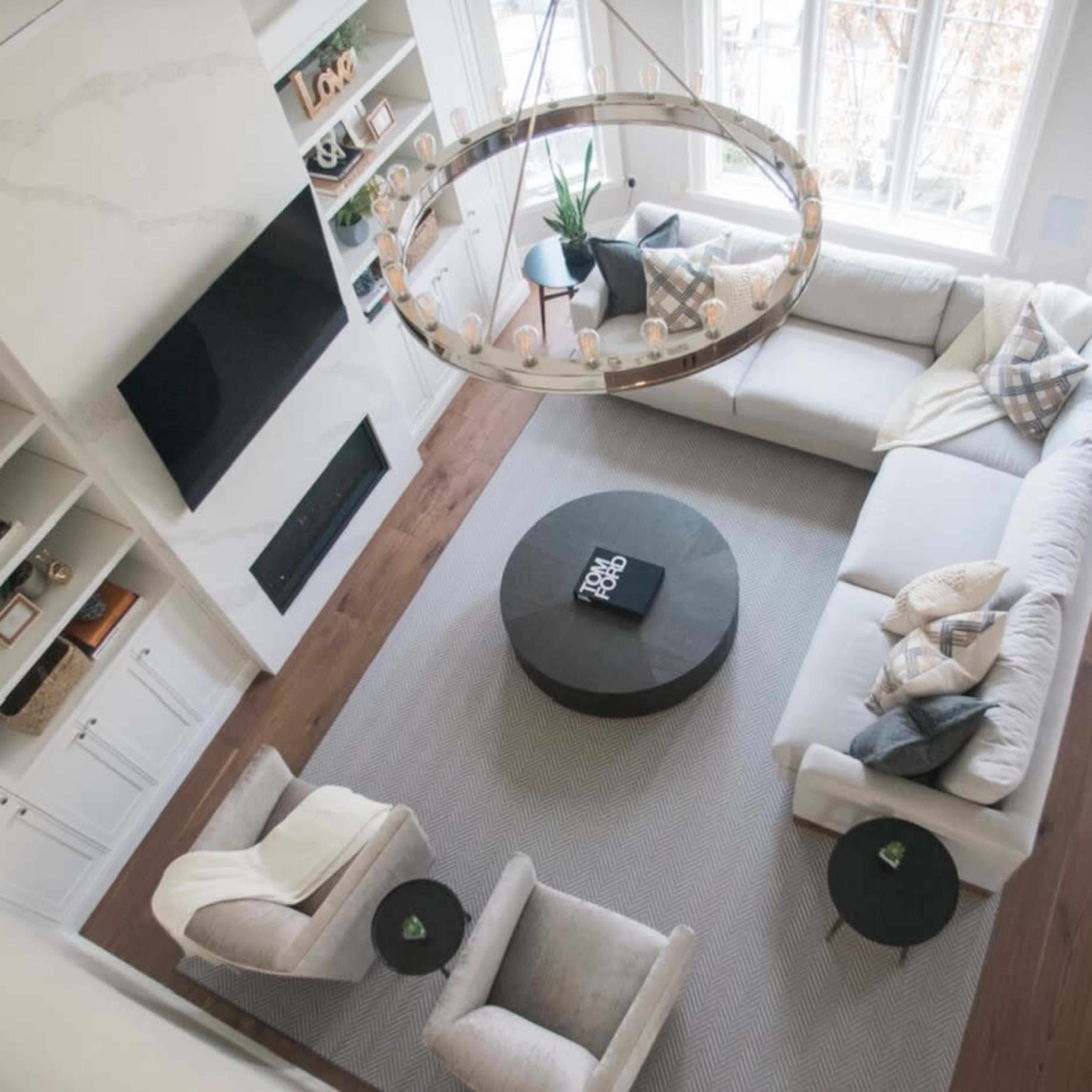 A modern living room with a large circular chandelier, light-colored furniture, and a central coffee table on a neutral rug, surrounded by large windows.