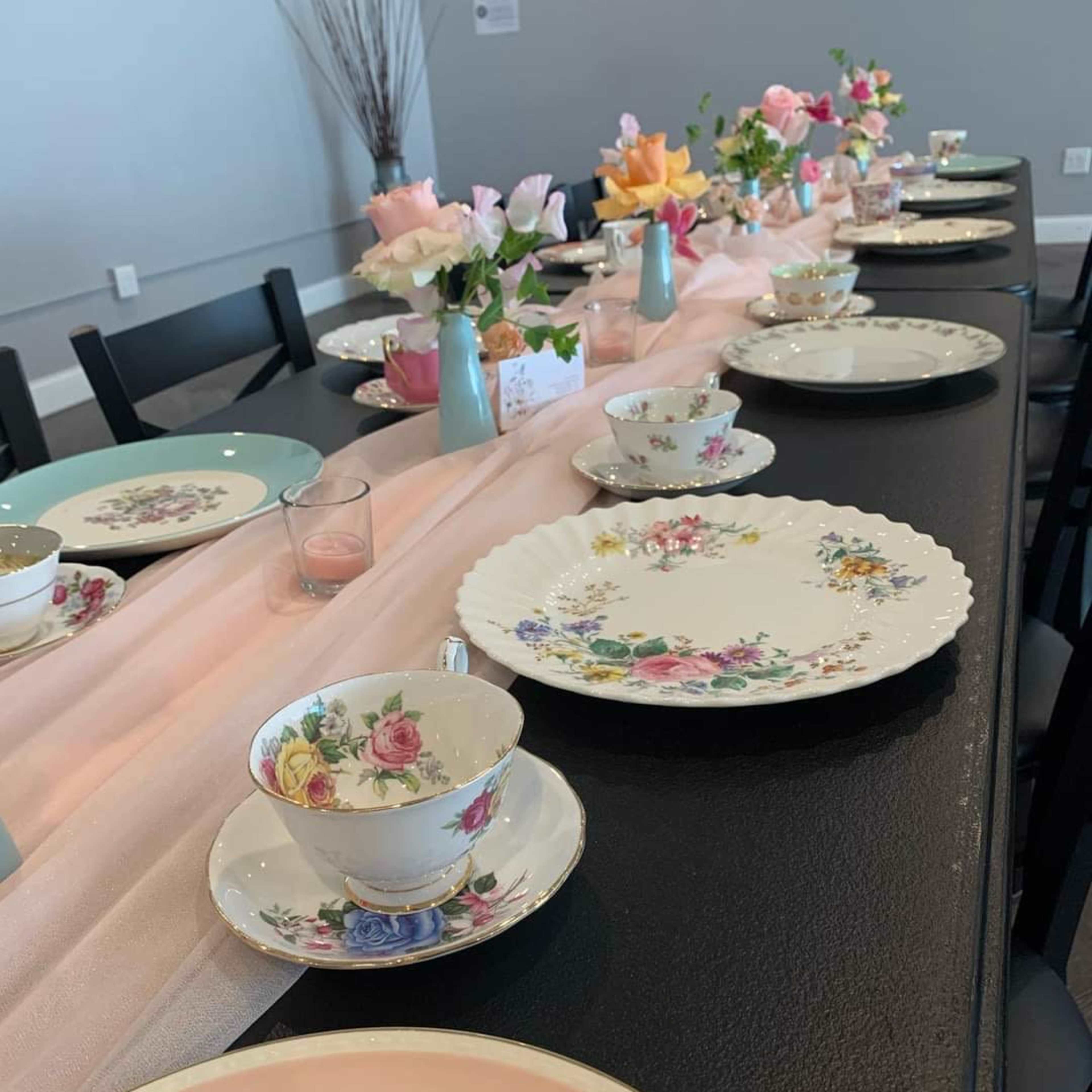A long dining table is set with a variety of floral-patterned china, including cups and plates, surrounded by small vases of flowers.