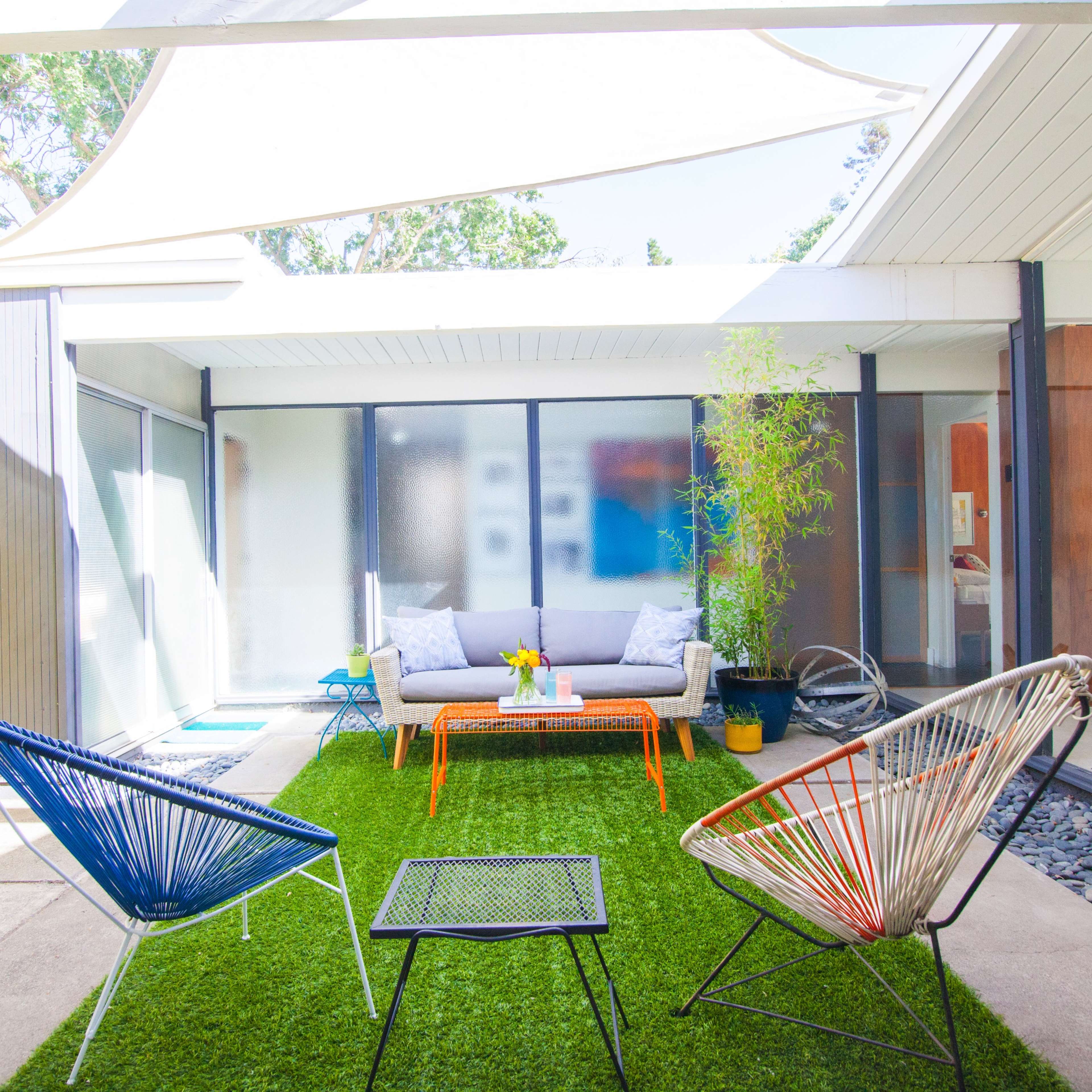 A bright outdoor living space features colorful chairs, a low table, and greenery, surrounded by modern glass walls.