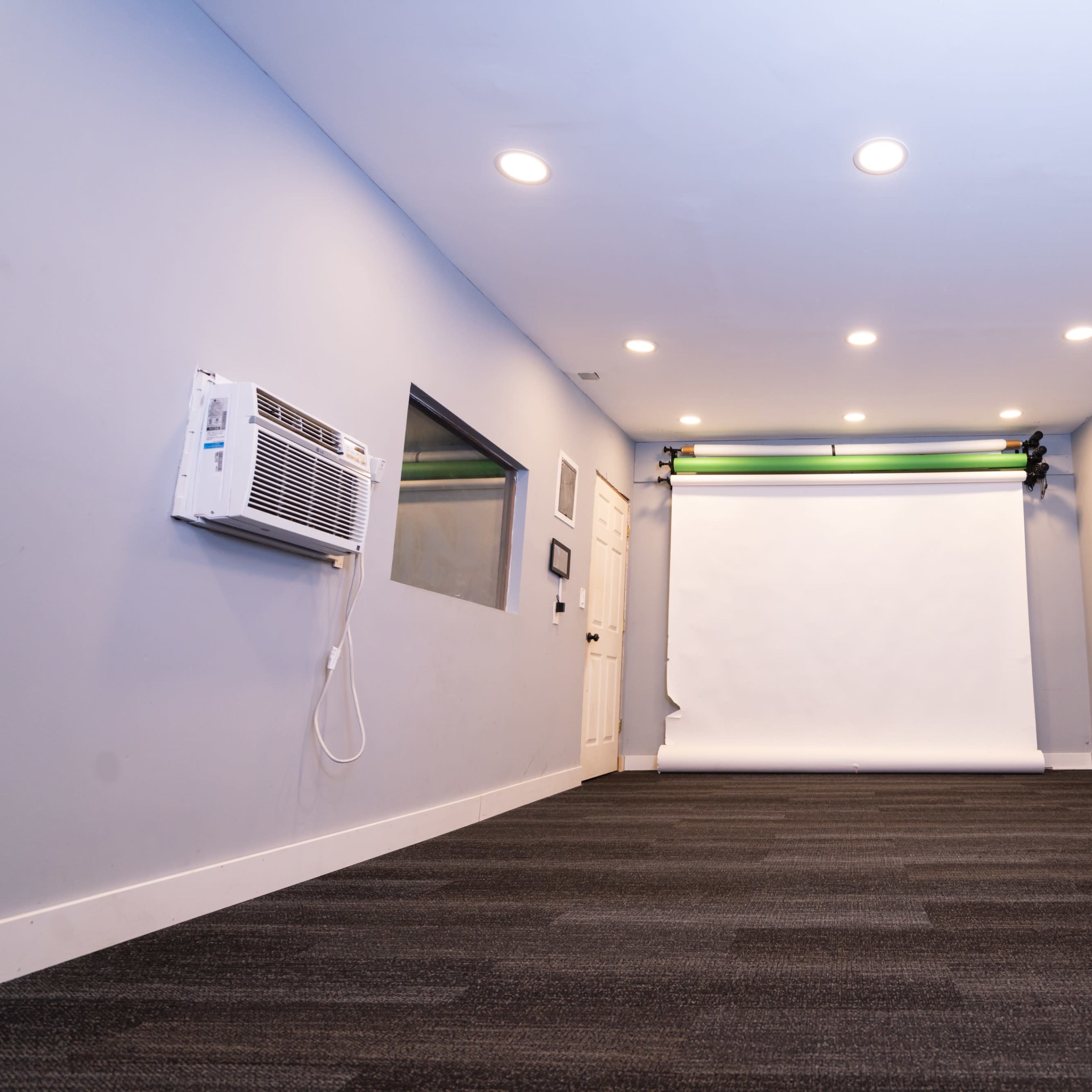 A minimalist studio space features a white backdrop, several chairs along one side, and an air conditioning unit on the wall.