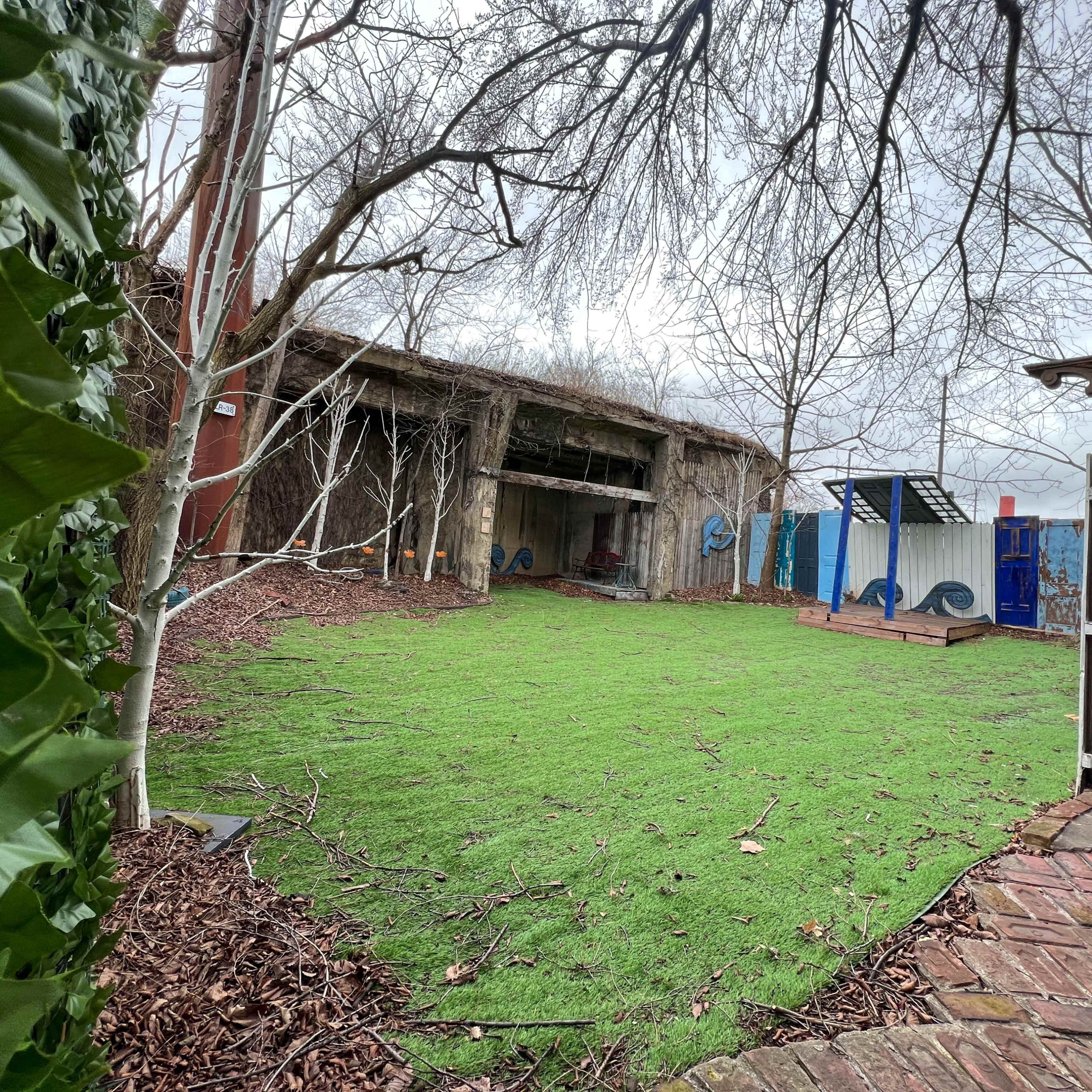 A grassy backyard area bordered by a wooden structure and trees, with a paved path on one side.