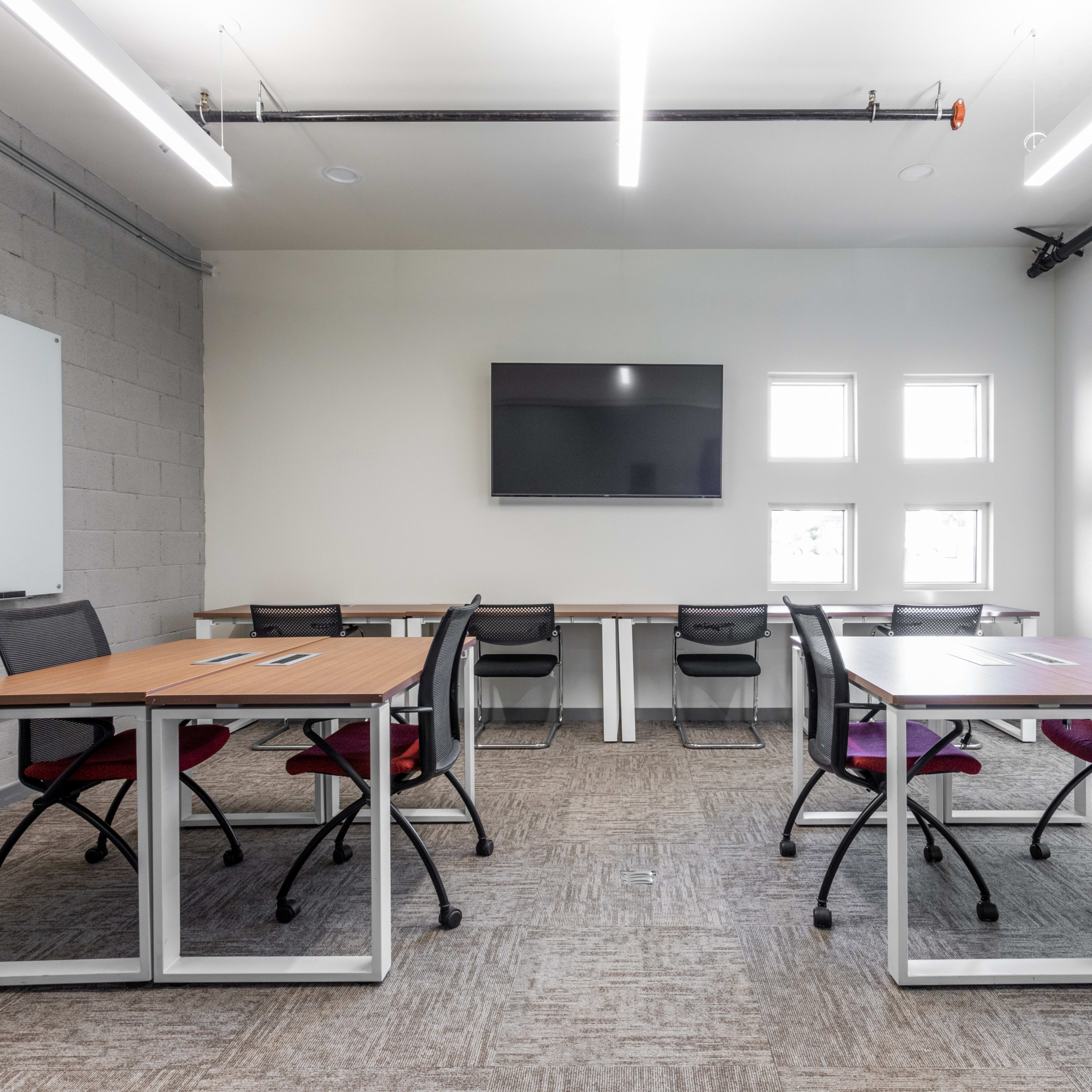 A modern office space featuring two sets of desks with chairs, a television mounted on the wall, and several windows.