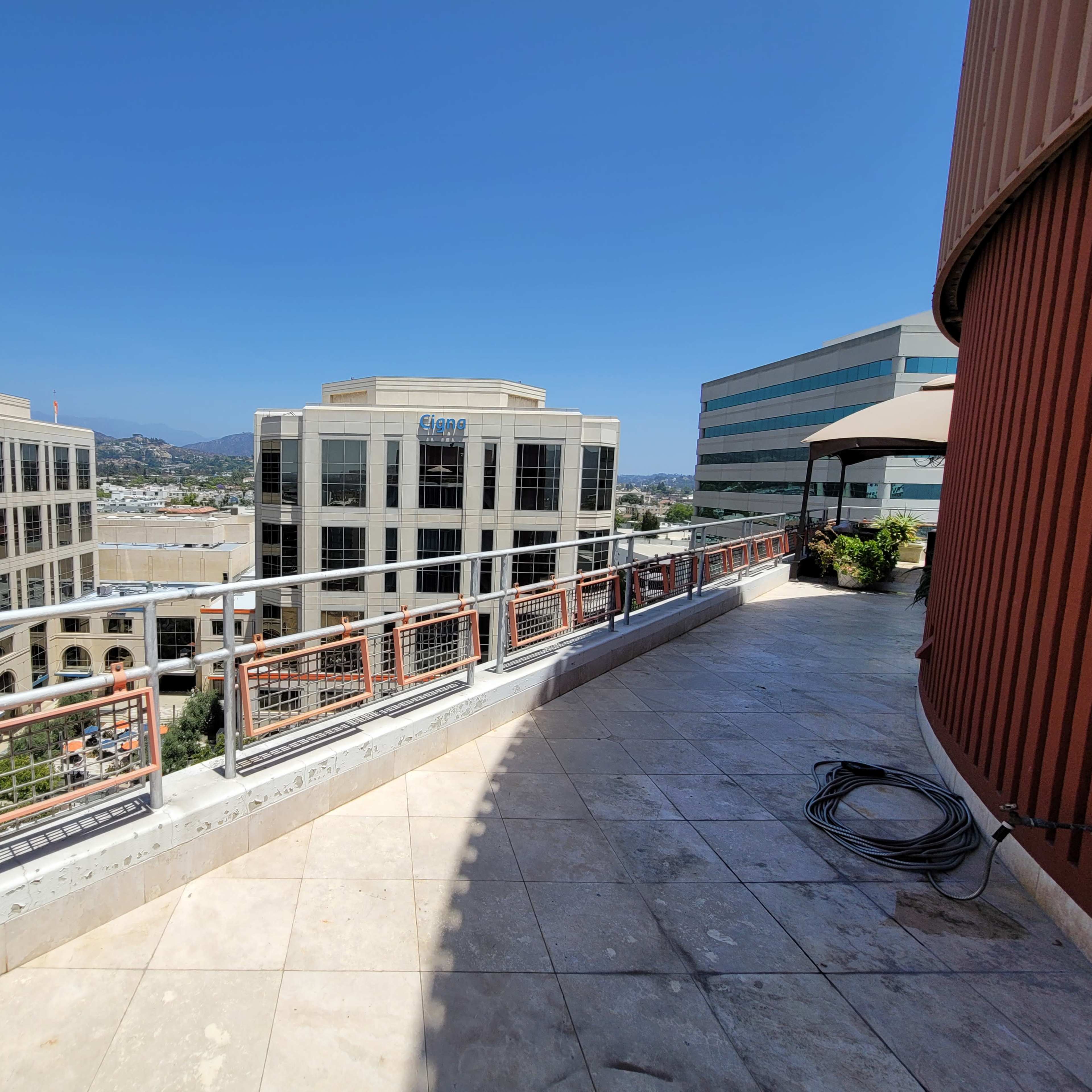 A sunlit balcony with tile flooring and a railing overlooking a row of commercial buildings and a clear blue sky.