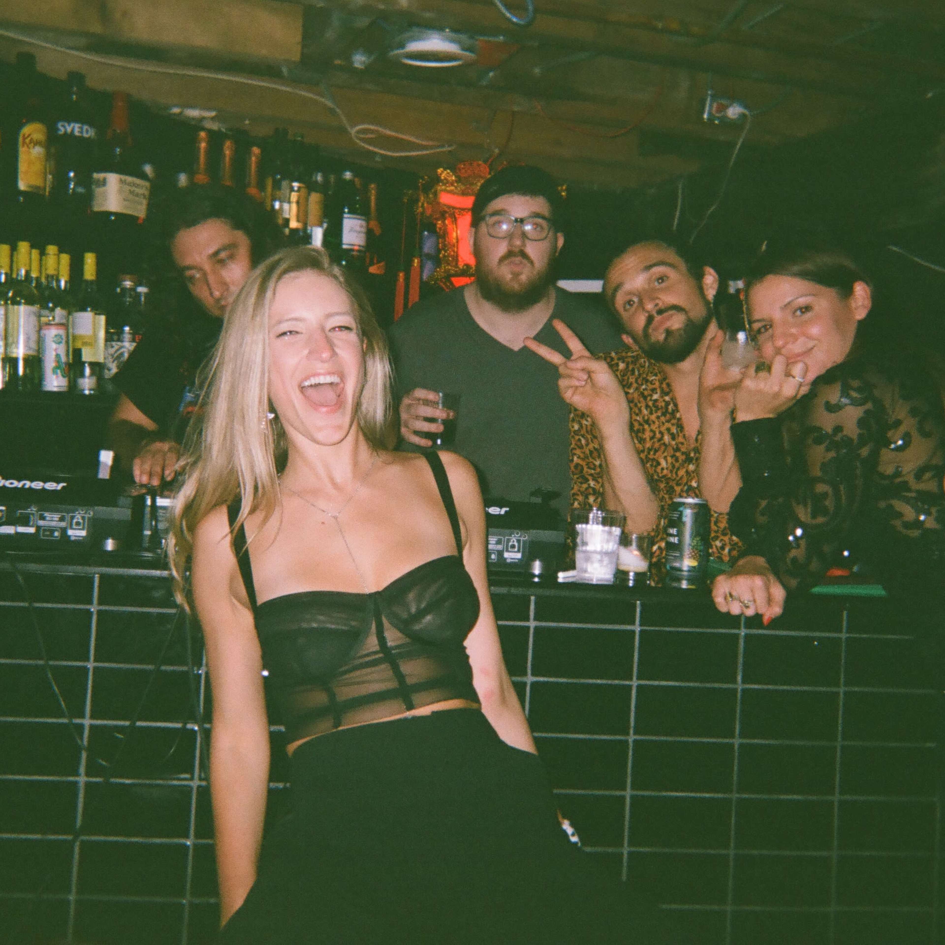 A woman poses playfully in front of a bar with five individuals behind her, surrounded by bottles and drinks.
