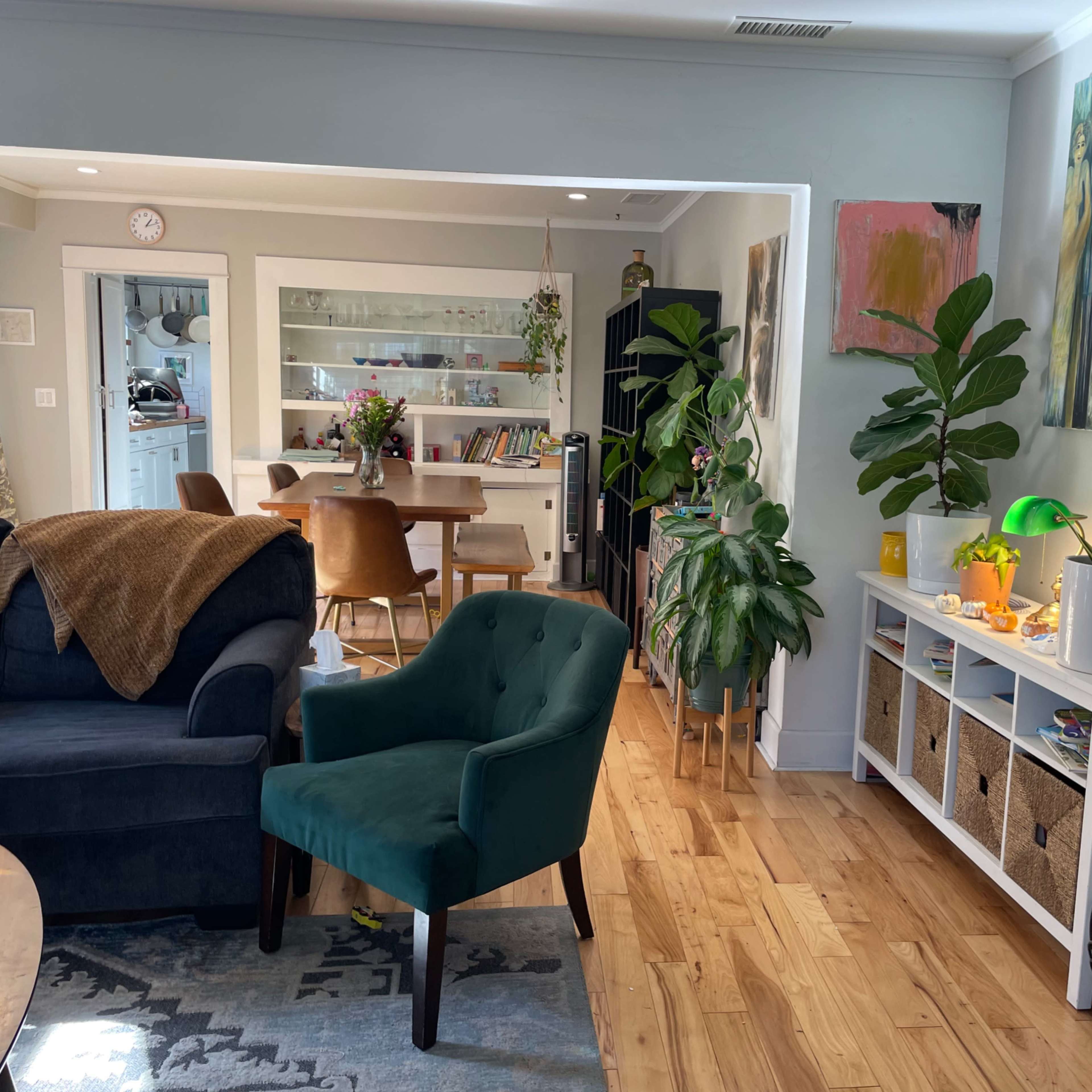 A living room connected to a dining area, featuring a blue sofa, a green chair, potted plants, and a white storage unit filled with baskets.