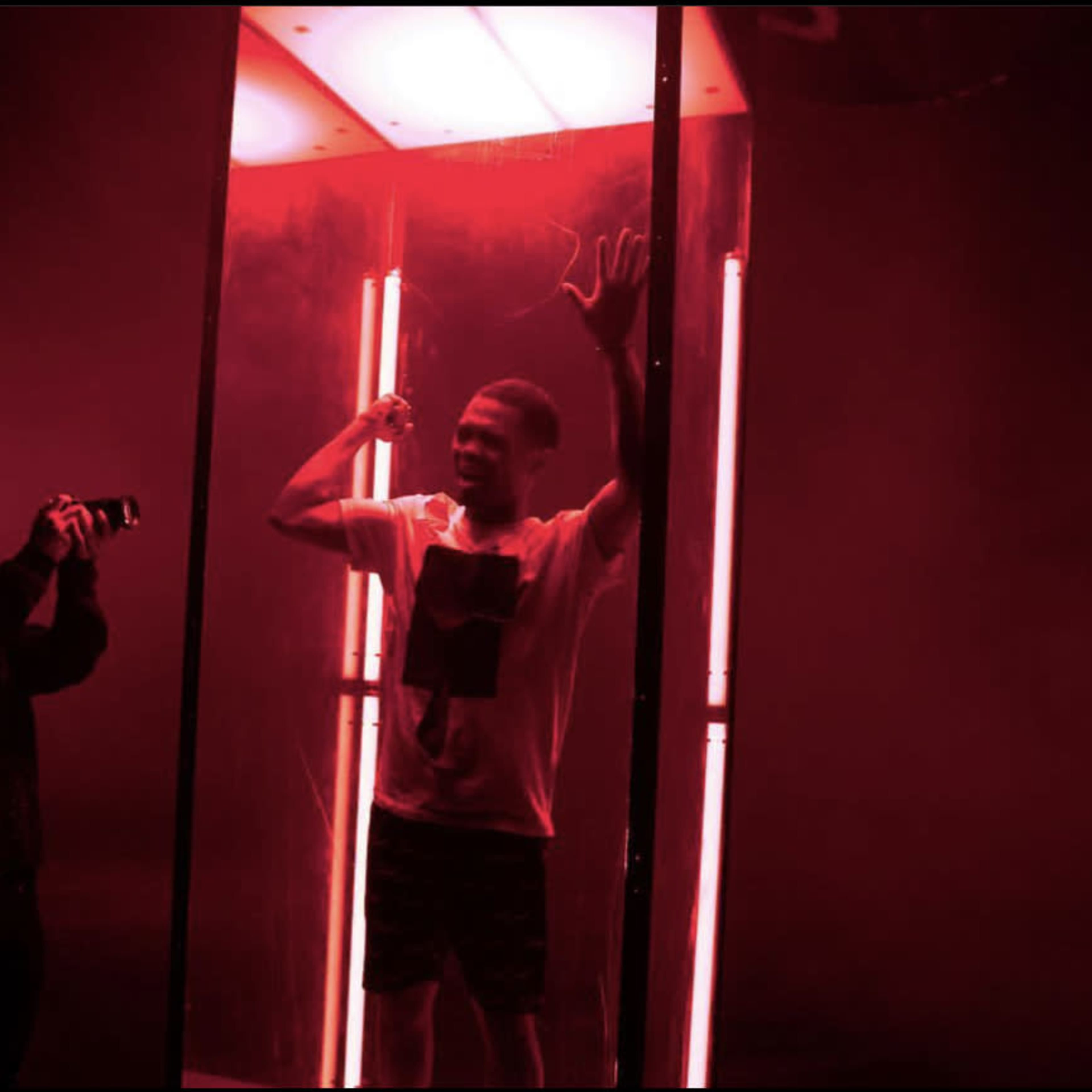 A person holds a camera while another stands inside a transparent box illuminated by red light.