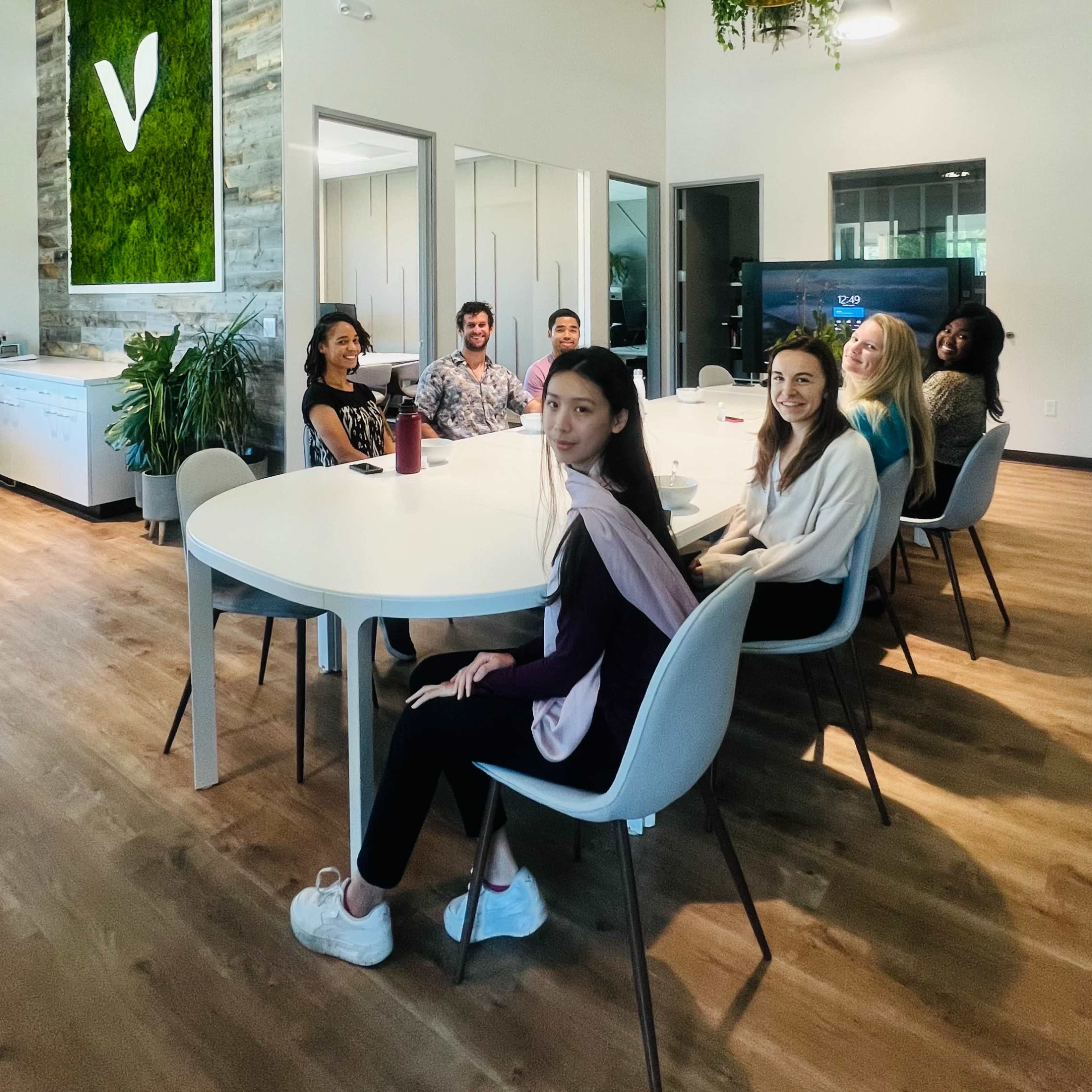 A group of people is sitting around a large, round table in a well-lit modern office with greenery on the walls and various plants around the room.