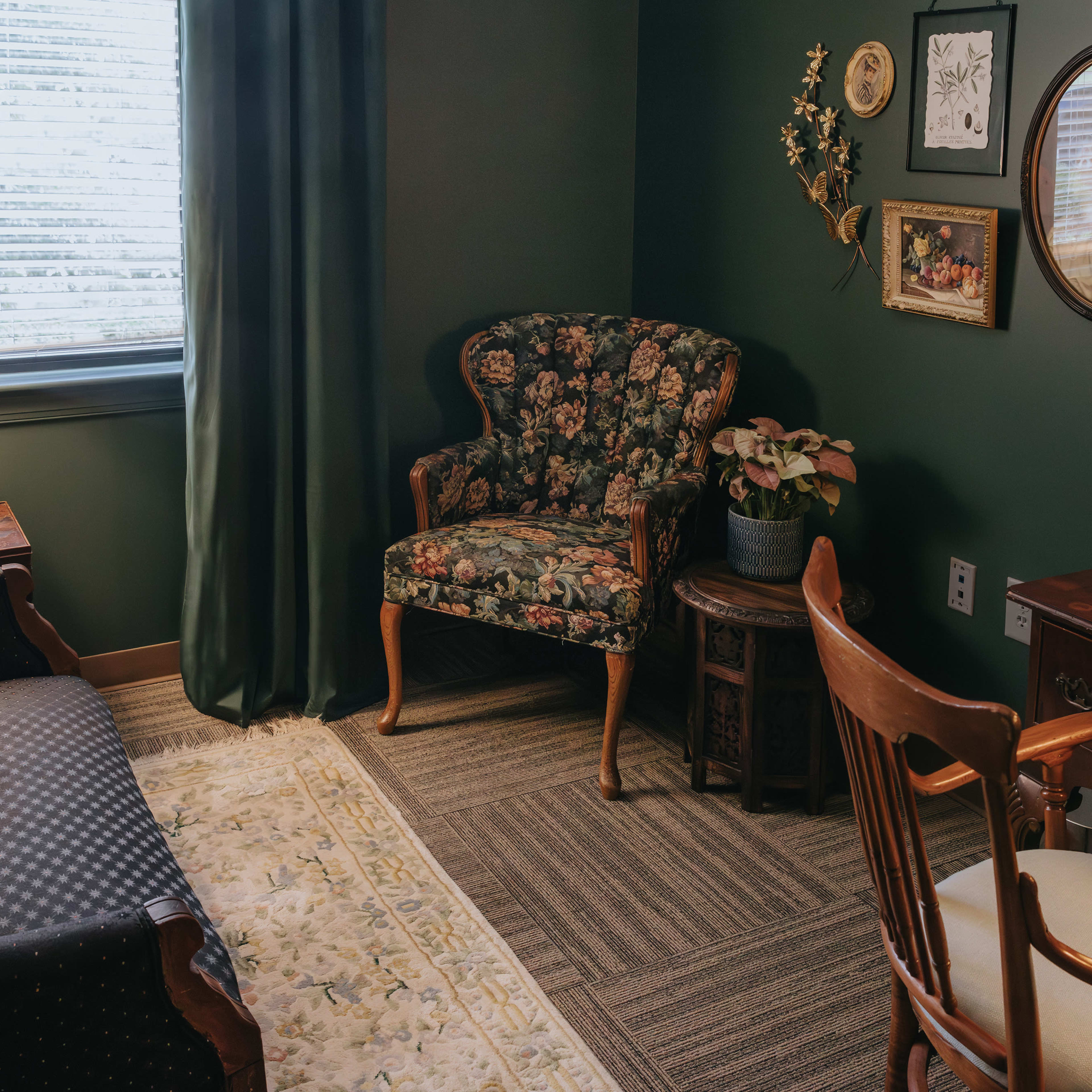 A cozy room with a floral armchair, a small wooden table, a mirror, and a vintage-style lamp beside a sofa on a patterned rug.