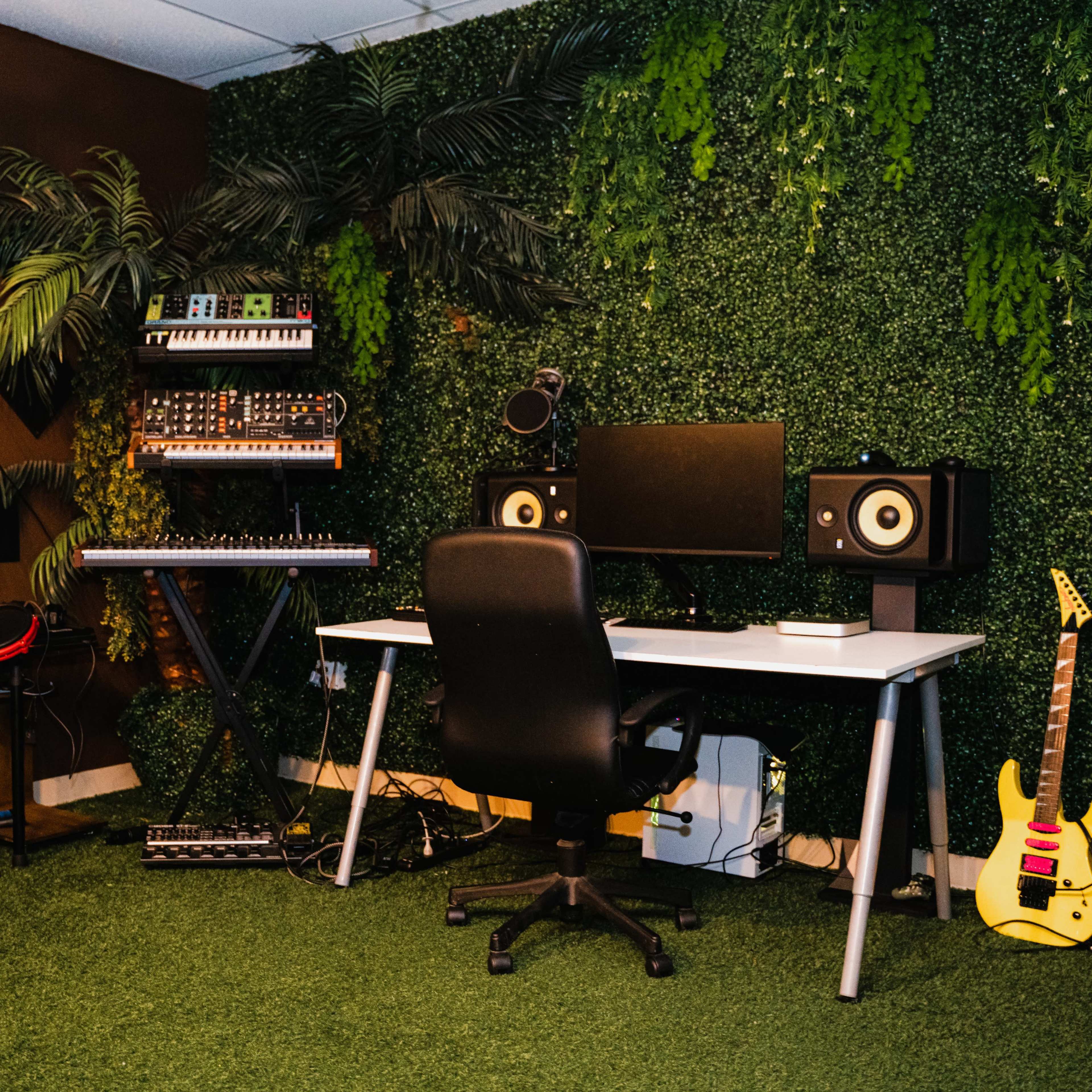 A music production setup features a desk with dual monitors, speakers, a keyboard, and a digital drum set in a room adorned with artificial greenery.