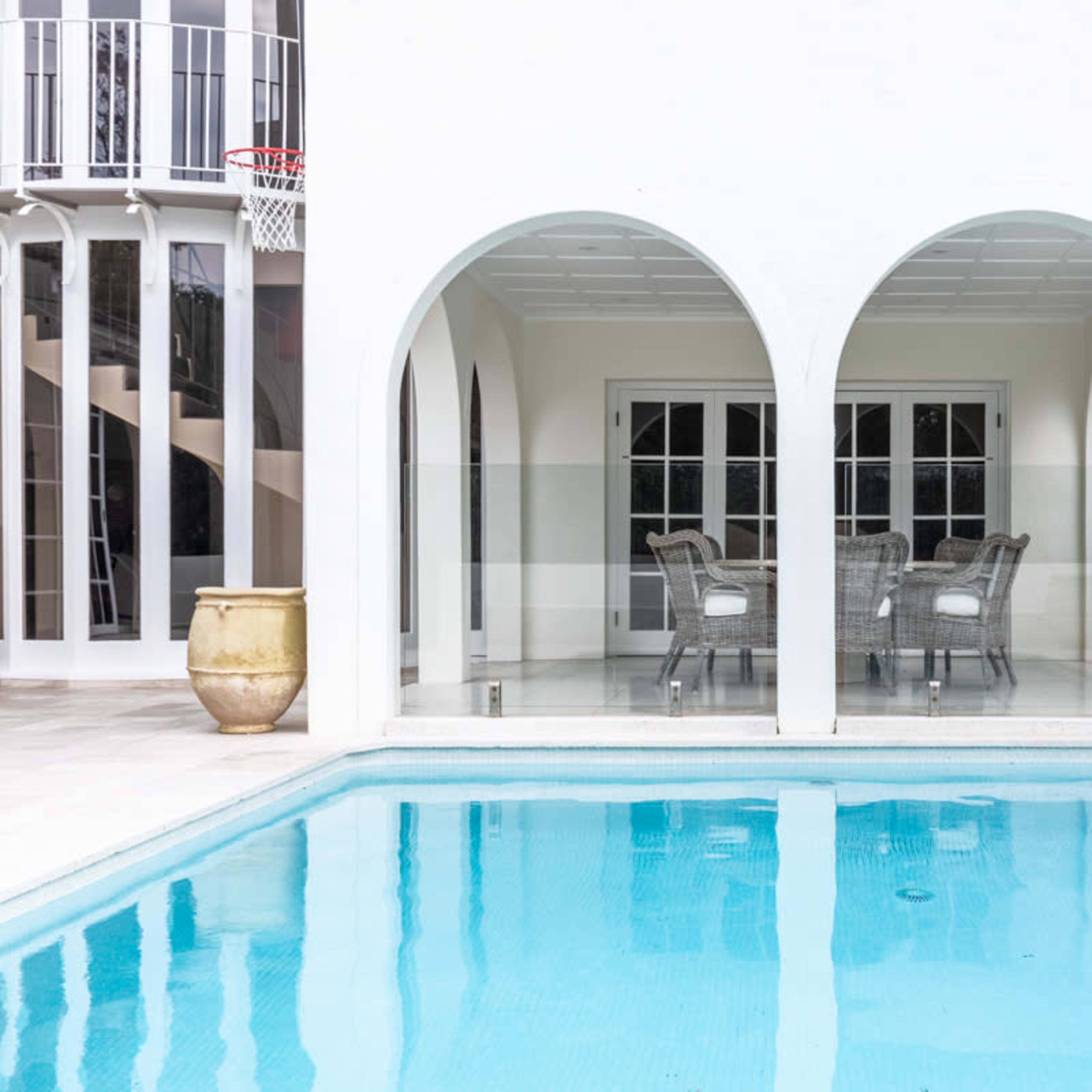 A swimming pool bordered by a white building featuring arched openings that lead to a glass-enclosed dining area.
