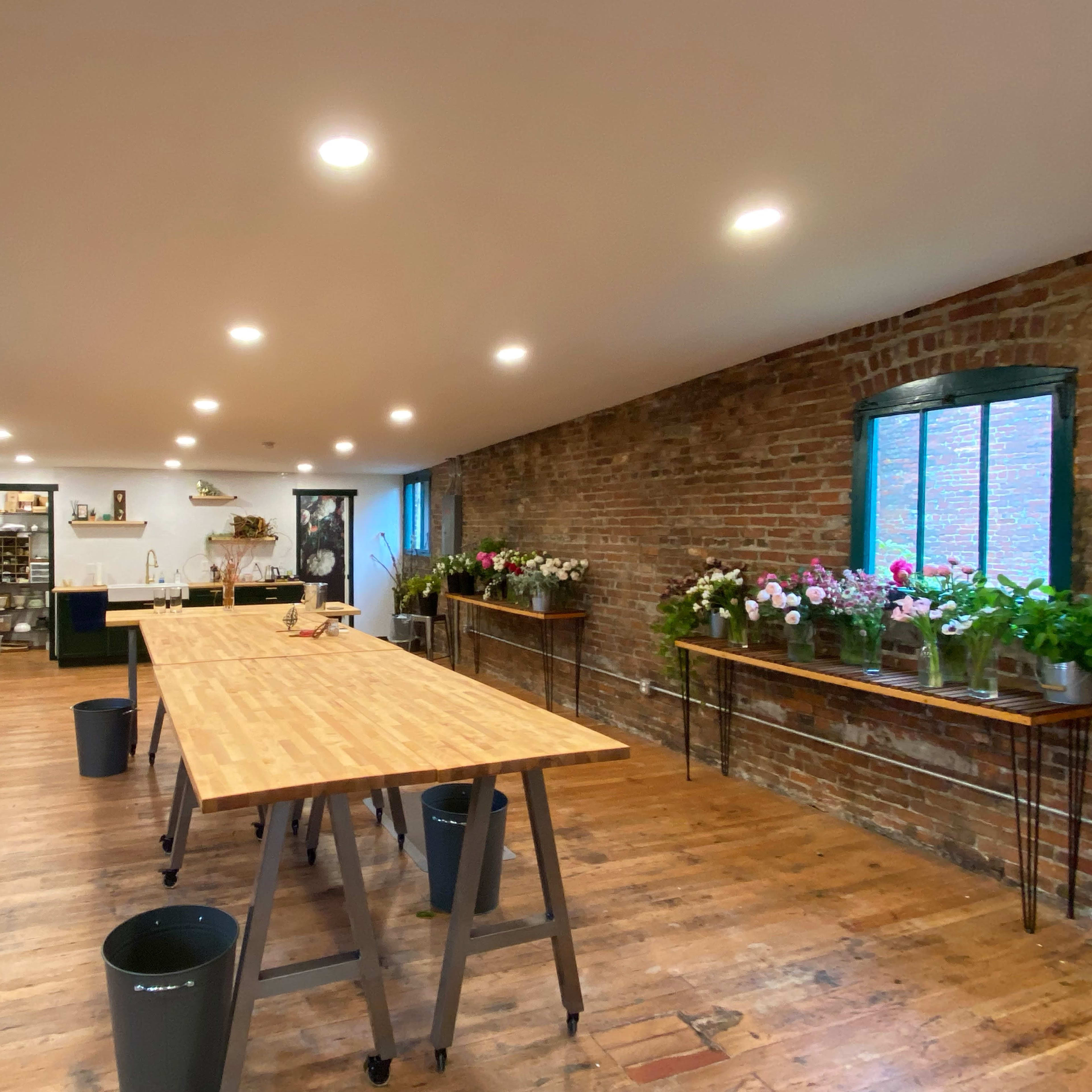 A spacious floral workshop featuring wooden tables, exposed brick walls, and various flower arrangements near the windows.