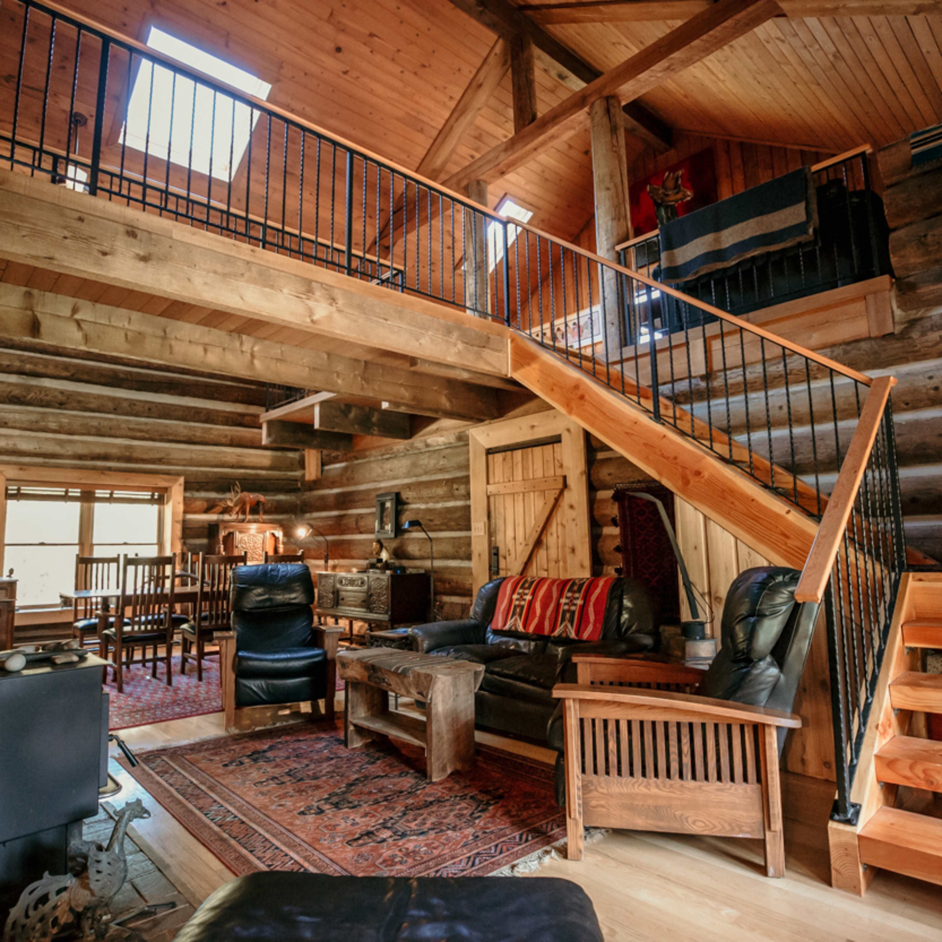 A rustic log cabin interior with a wooden staircase, leather seating, and a dining area visible in the background.